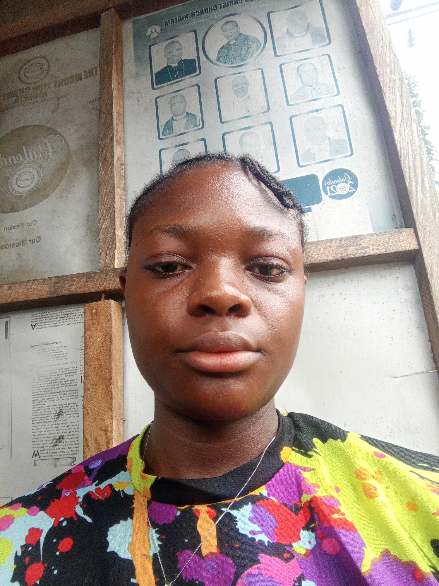 A young Black woman is captured in a close-up portrait, likely a selfie, in Mbribit, Nigeria. She looks directly at the camera with a neutral expression. Her dark hair is styled in braids and pulled back. She wears a vibrant, multi-colored top with a painterly, abstract design featuring splashes of purple, yellow, red, light blue, and black. A thin silver necklace is visible around her neck.
The background consists of a wall or notice board framed by raw wooden beams. This board displays several notices and photographs, many of which appear inverted or reversed. On the right, a grid of faded black and white portrait photographs, possibly of clergy members, is visible. Above these photos, the inverted text "CHRIST CHURCH NIGERIA" is discernible. Below them, a circular logo contains the word "JOSI" and what might be a year like "2023" or "2025". On the left side, another inverted circular logo, possibly related to "Calvary" or "THE HOLY VISION CHURCH" (from "THE WOOHL SION CHEIRA"), is visible alongside more inverted and garbled text fragments, including phrases like "Our Mission" (from "marw nozziM bhow") and other document excerpts.
The setting appears to be an indoor or sheltered area, possibly a community hall or a church building, suggested by the religious references in the background text and images. The lighting is soft and even, indicating daytime. No specific activity beyond posing for the photograph is depicted.