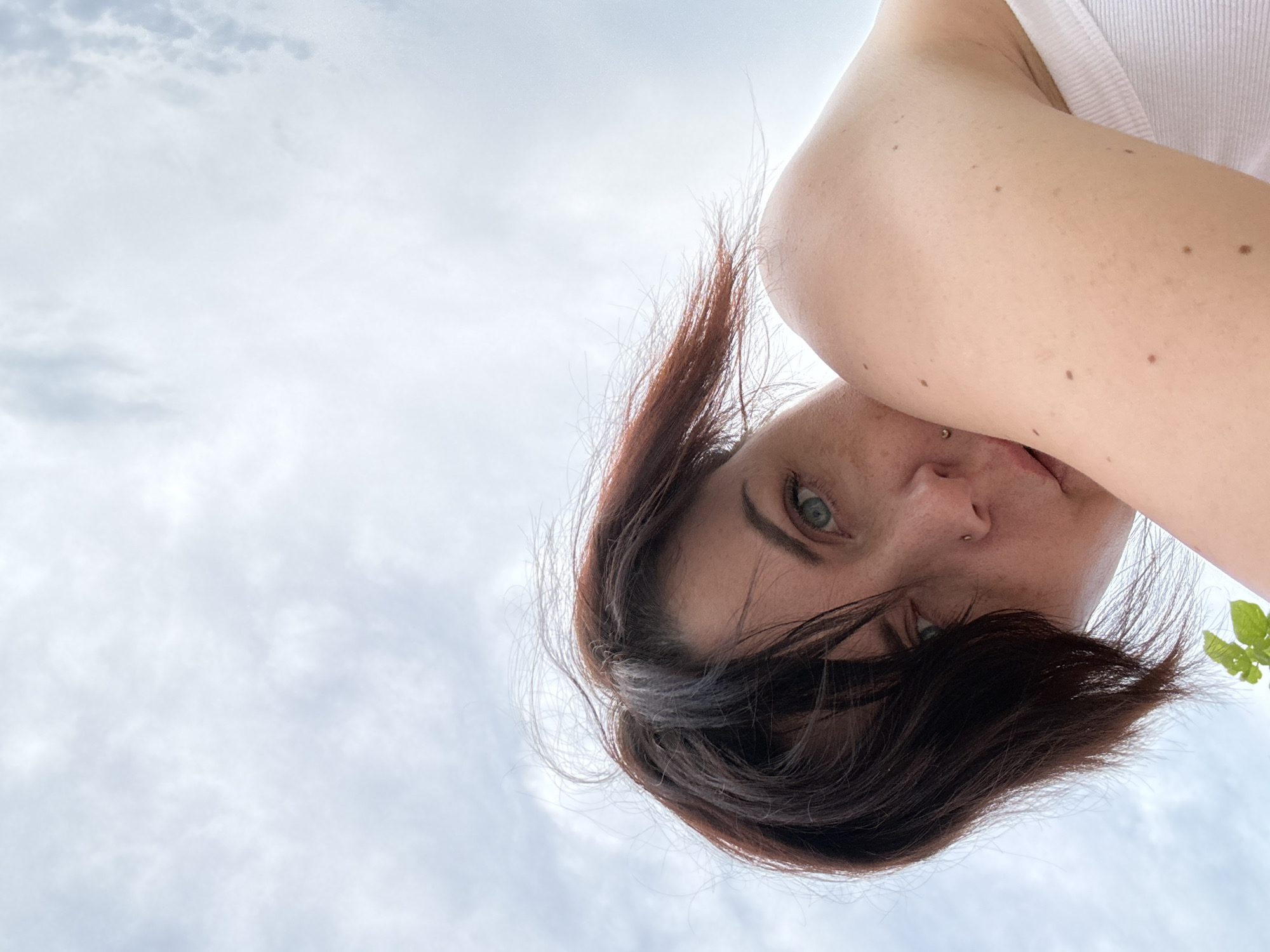The image is a low-angle, upside-down selfie taken outdoors, likely in a rural or natural setting given the context of Berezyna, Ukraine. The primary subject is a person, their face angled towards the camera and partially obscured by their own dark hair. They have piercing blue-green eyes, dark eyebrows, and a small nose stud. Their skin is fair and appears to have several moles. The person is wearing a white tank top.
The background consists of an overcast sky with soft, diffused clouds, suggesting a daytime setting that is either cloudy or perhaps early morning or late evening. There are no clear architectural or man-made structures visible, and the only hint of greenery is a small leaf in the upper right corner of the frame, suggesting vegetation nearby. The overall mood is somewhat introspective or perhaps candid, with the low angle and obscured view adding a sense of immediacy and informality. There is no visible text in the image.
