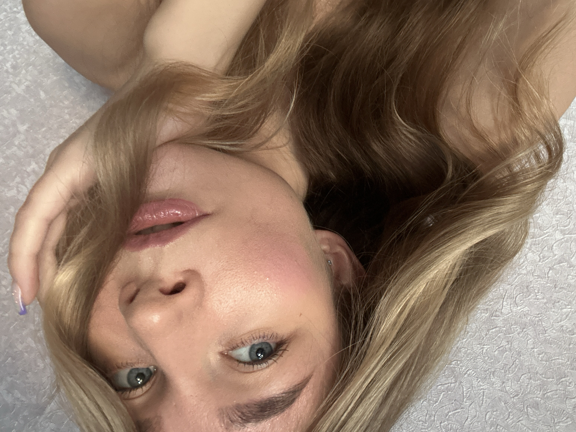 A close-up, rotated portrait shows a young woman's face, with her head tilted to the left and her right hand resting near her temple, her fingers brushing against her blonde hair. Her blue eyes are looking upwards, and her lips are slightly parted, revealing a hint of her tongue. She is wearing subtle makeup, with a pink blush on her cheeks and a glossy pink lipstick. A small, silver hoop earring is visible in her left ear. Her blonde hair is long and wavy, framing her face and falling over her shoulder. The background is a textured, light-colored surface, possibly a wall or fabric. The lighting is soft and diffused, highlighting the contours of her face and hair. The image has a personal and intimate feel, suggesting a candid moment.
