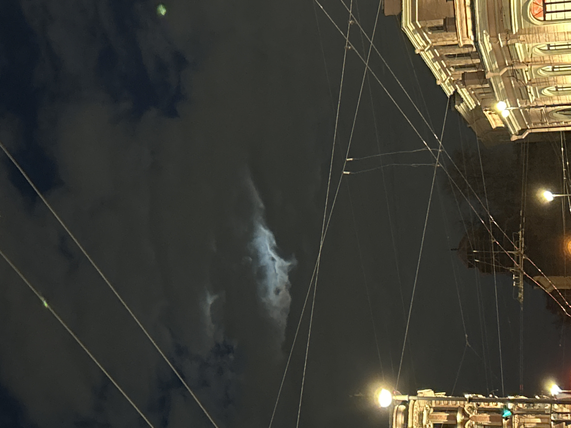 The image captures a nighttime urban scene, appearing to be an upward-looking shot originally taken in portrait orientation and rotated 90 degrees clockwise. The dark sky, largely covered by scattered clouds, dominates the frame. Through a break in the clouds in the mid-left, a bright, glowing moon is partially visible.
Numerous thin, light-colored wires crisscross the entire sky, running diagonally and horizontally, suggesting a network of overhead power lines, trolleybus, or tram wires. On the right side of the frame, the ornate facade of a building with classical architectural elements is prominently illuminated. Details such as arched windows, cornices, and columns are visible under a warm light. Below and to the right, several bright streetlights or building lights cast a soft glow. A faint, small green light or lens flare is visible in the upper-left corner of the sky.
No people are visible in the scene, and there is no discernible text. The image conveys the atmosphere of a cool, cloudy night in an older European city, aligning with the provided context of Saint Petersburg, Russia.