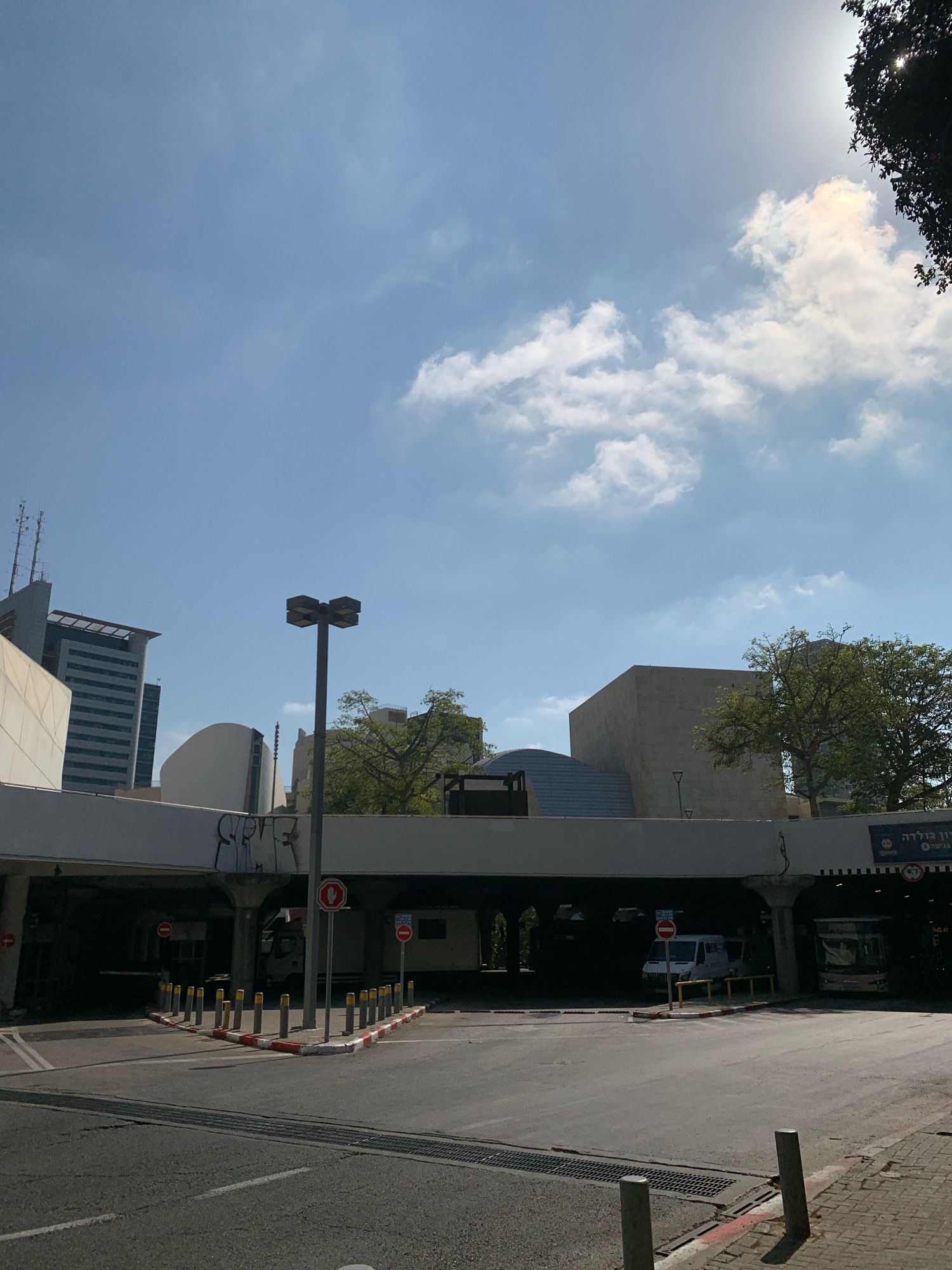 The image shows a street scene in Tel Aviv, Israel, with an overpass and modern buildings. The overpass has a white railing and graffiti on its underside. There are traffic signs, bollards, and a street light. A white van and a bus are visible under the overpass. In the background, tall buildings with antennas and trees are seen under a blue sky with scattered clouds and bright sunlight. A sign with Hebrew text is visible on the right side of the overpass. The ground is paved and appears to be a road or parking area with a drainage grate.