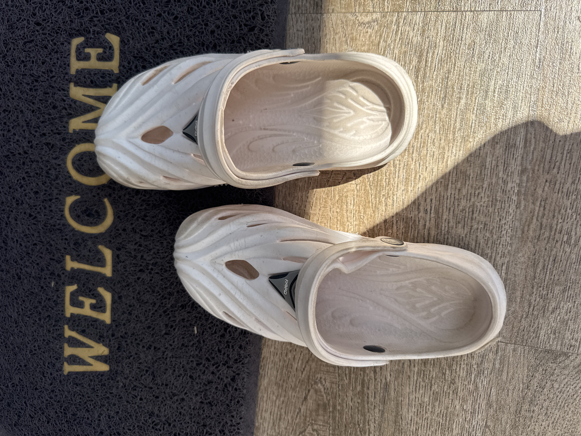 The image shows a pair of white clogs on a surface that is part "welcome" mat (black with gold lettering) and part light wood-grain flooring. The flooring appears to be an interior surface, possibly in Kuta, Indonesia, but there are no identifying features to confirm this. There are no people visible.