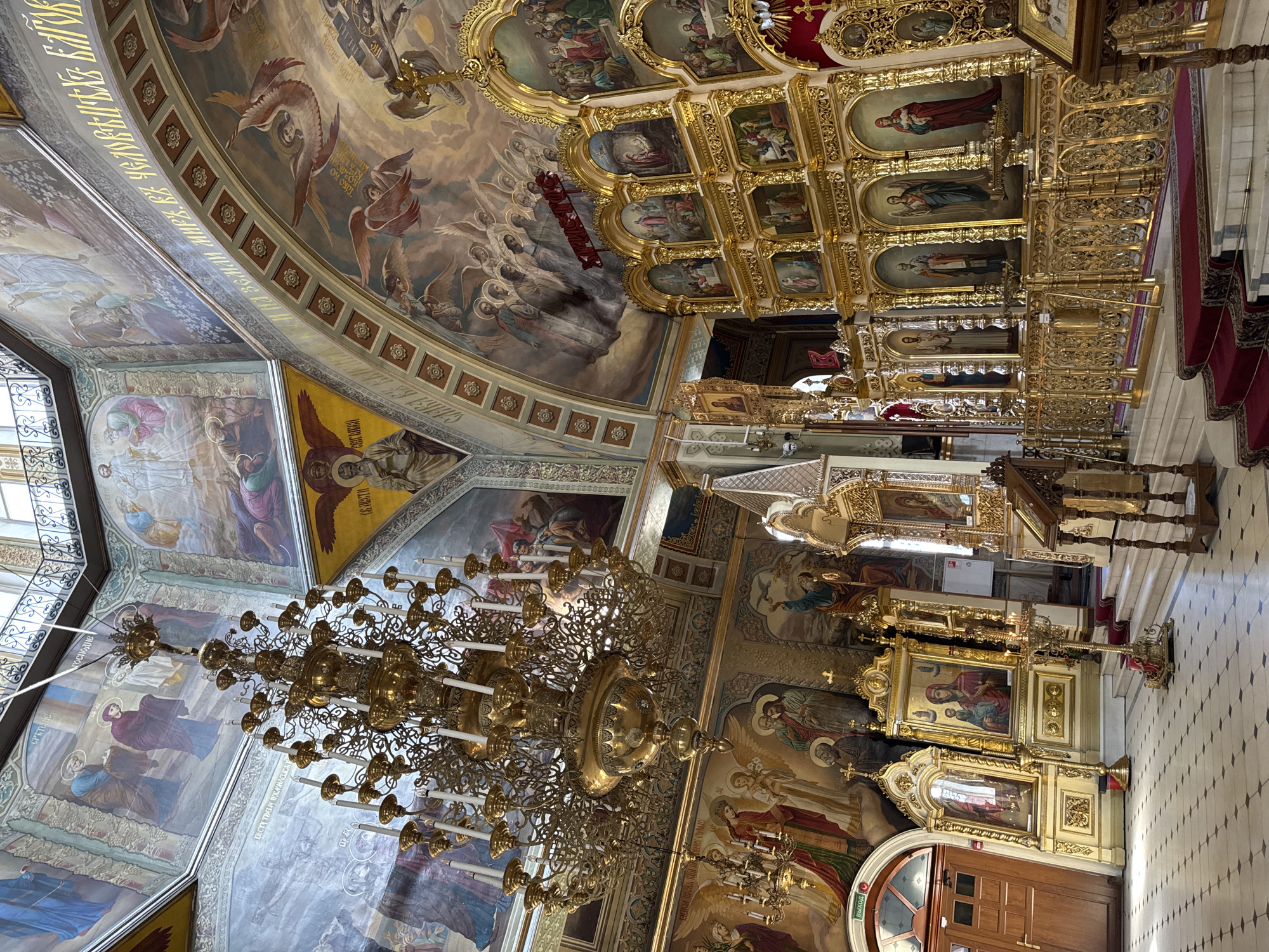 This is a high-angle, full shot of the interior of an ornate Orthodox church, likely in Eastern Europe, featuring rich Byzantine-style iconography and gilded architectural elements. The dominant feature is a large, multi-tiered brass chandelier with numerous candle holders, suspended from the vaulted ceiling. The walls and ceiling are covered in vibrant frescoes depicting religious scenes and figures, rendered in a traditional Eastern Orthodox style with rich colors and detailed figures.
The foreground is dominated by a massive, gilded iconostasis, a screen separating the nave from the sanctuary, adorned with multiple panels of icons. These icons are framed in gold and depict various saints and biblical narratives. The iconostasis is backed by ornate gold railings, and in front of it, red carpets cover tiered white marble steps. To the left, a large wooden door with arched glass panes stands open, revealing a glimpse of the exterior.
Other elements in the scene include smaller chandeliers, various religious icons displayed on pedestals, and wooden lecterns. The floor is tiled in a black and white checkerboard pattern. The lighting is a mix of natural light filtering in from high windows and the artificial light from the candles in the chandeliers. The overall impression is one of opulence, reverence, and artistic grandeur, typical of a well-preserved historical Orthodox church. There are no people visible in the scene.