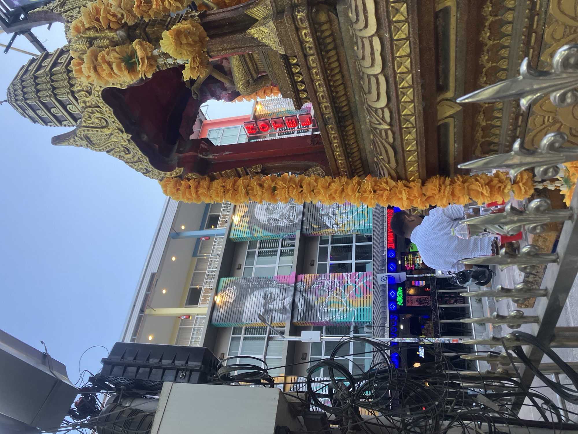 An outdoor urban scene in บ้านบางรักษ์, Thailand, is captured from a low angle, looking upwards. In the immediate foreground and extending through the frame is a highly ornate, golden structure, possibly part of a shrine, float, or building facade. It is richly decorated with intricate carvings and adorned with bright yellow flower garlands, likely marigolds. A silver metal railing with spiked accents is also prominent in the foreground.
Beyond this structure, a person wearing a white polka-dot shirt is partially visible, seated, suggesting they are at a street-side establishment. Behind them, multiple-story buildings line the street. One building features large, striking monochromatic murals of faces integrated with colorful striped patterns across its facade. Various commercial signs are visible on the buildings, including a red neon sign displaying "FOOD," and blue neon signs that read "JINN INN," "AIRCON," and partially "SOON." A smaller sign with "TIMMY WOODS" is also discernible.
Above the street level, a dense web of black electrical wires and junction boxes stretches across the frame, characteristic of urban infrastructure in many Southeast Asian cities. The sky is clear and light blue, indicating daytime, although some neon signs are illuminated. The overall impression is a bustling, culturally rich street scene with a blend of traditional ornamentation and modern urban elements.