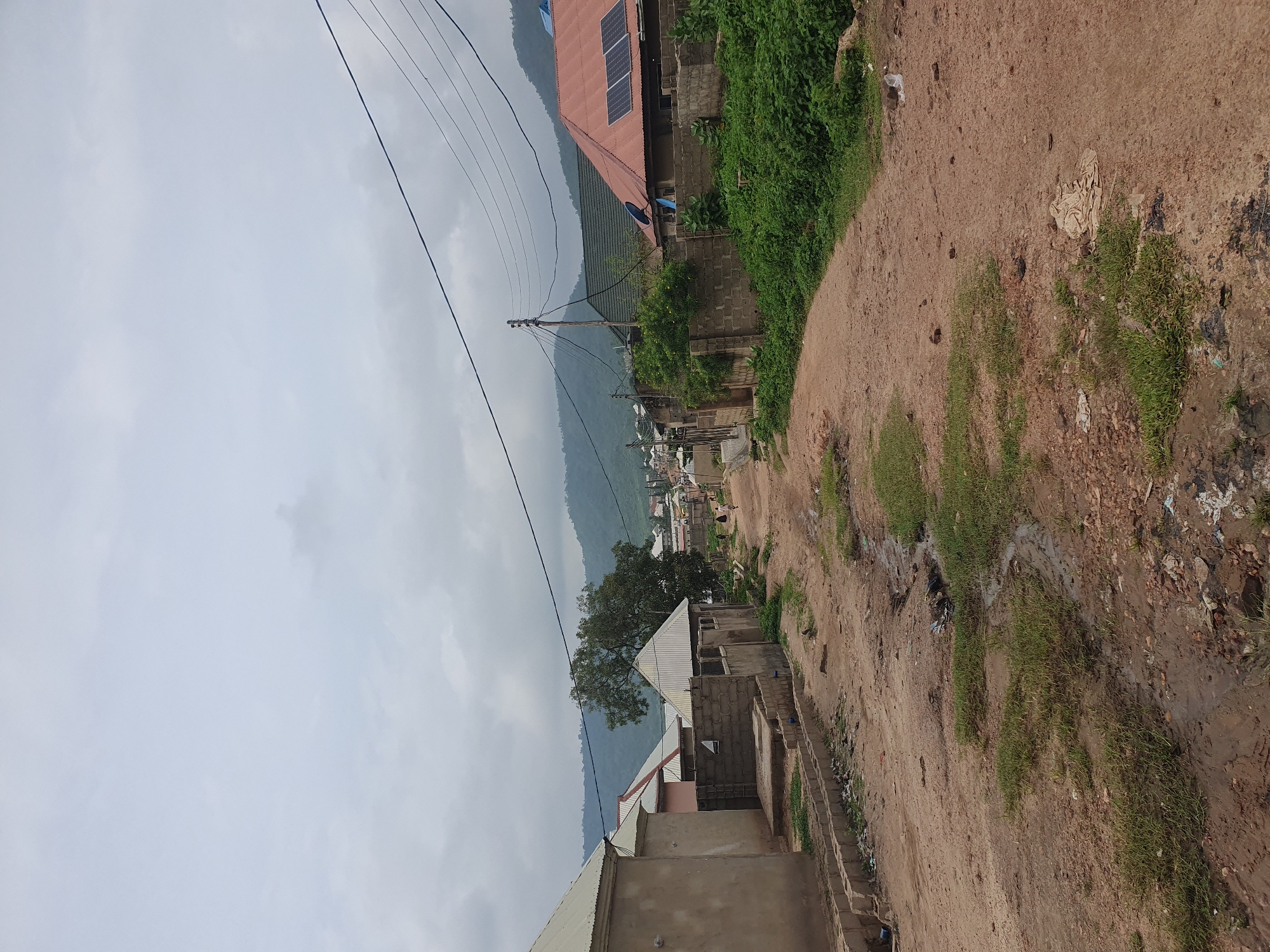The image depicts a street scene in Lokoja, Nigeria, characterized by unpaved roads and modest residential structures. The road surface is composed of reddish-brown dirt, with patches of sparse grass and evidence of water accumulation, suggesting recent rainfall or poor drainage. Debris, including plastic waste and discarded items, is visible along the roadside and in the drainage channels.
Several buildings are present, varying in their stage of construction and materials. Some structures have finished roofs, including corrugated metal and a tiled roof with solar panels. Others are partially constructed, with exposed brickwork. Fencing, constructed from concrete blocks, encloses some properties.
Vegetation is present, with overgrown green bushes lining parts of the street and a mature tree visible further down the road. In the background, rolling hills are discernible under a cloudy sky. Power lines and a utility pole are visible, indicating the presence of electrical infrastructure. A few individuals are walking along the road in the distance, suggesting community activity. The overall impression is a rural or semi-urban residential area with developing infrastructure.