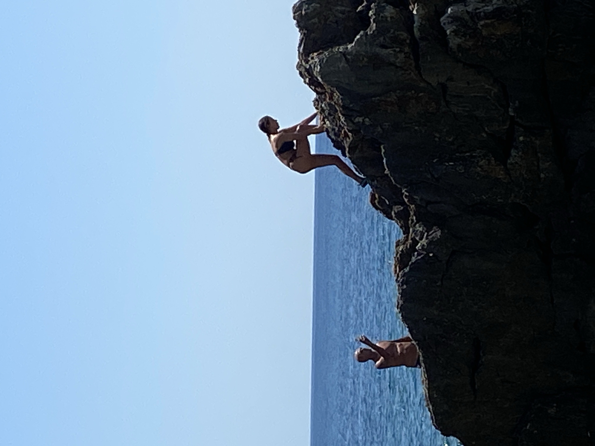 A medium shot captures two individuals on a rocky coastline with a clear blue sky and a deep blue sea as the backdrop. On the left, a bare-chested person stands on a rock, facing right with their left arm raised and hand extended as if pointing or holding something. They appear to be wearing dark briefs. To the right, a person in a bikini is actively climbing a large, dark rock formation. The person in the bikini is positioned higher up on the rock, with their body bent as they reach for a higher foothold. The rocky terrain is rough and textured, with shadows indicating strong sunlight. The sea stretches out behind the figures, appearing calm and vast. The overall scene suggests a summer day at the coast in Prijevor, Montenegro, with people enjoying the natural environment.