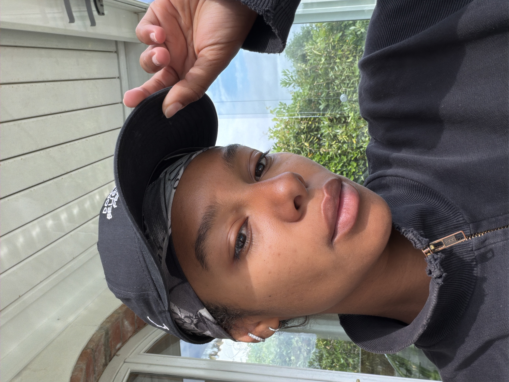 A person with dark skin is wearing a black baseball cap with white logos and a black zippered hooded sweatshirt. A gray patterned bandana is visible under the hat. The person is holding the brim of the cap with their hand. They have an earring in their left ear. The background appears to be a window with a view of trees and the sky. The location is El Mas Tomasí, Spain.