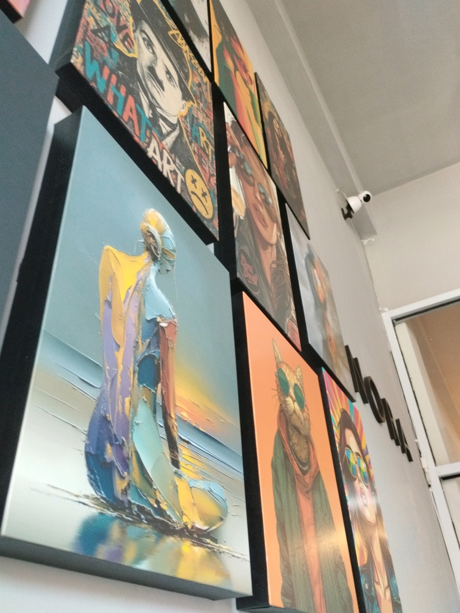 The image displays a wall adorned with several canvas paintings, viewed from a low angle looking upwards and to the right. The paintings are a mix of styles, including a portrait with graffiti-like elements, an abstract figurative piece in muted blues and yellows, a whimsical depiction of a cat wearing sunglasses, and another portrait of a woman with sunglasses. A white security camera is mounted on the wall above and to the right of the paintings. The overall impression is of an art display within an indoor space, possibly a gallery or a shop. The prompt requests information about the location in Gorontalo, Indonesia, but the image itself does not contain any visual cues that would indicate this specific geographical location.