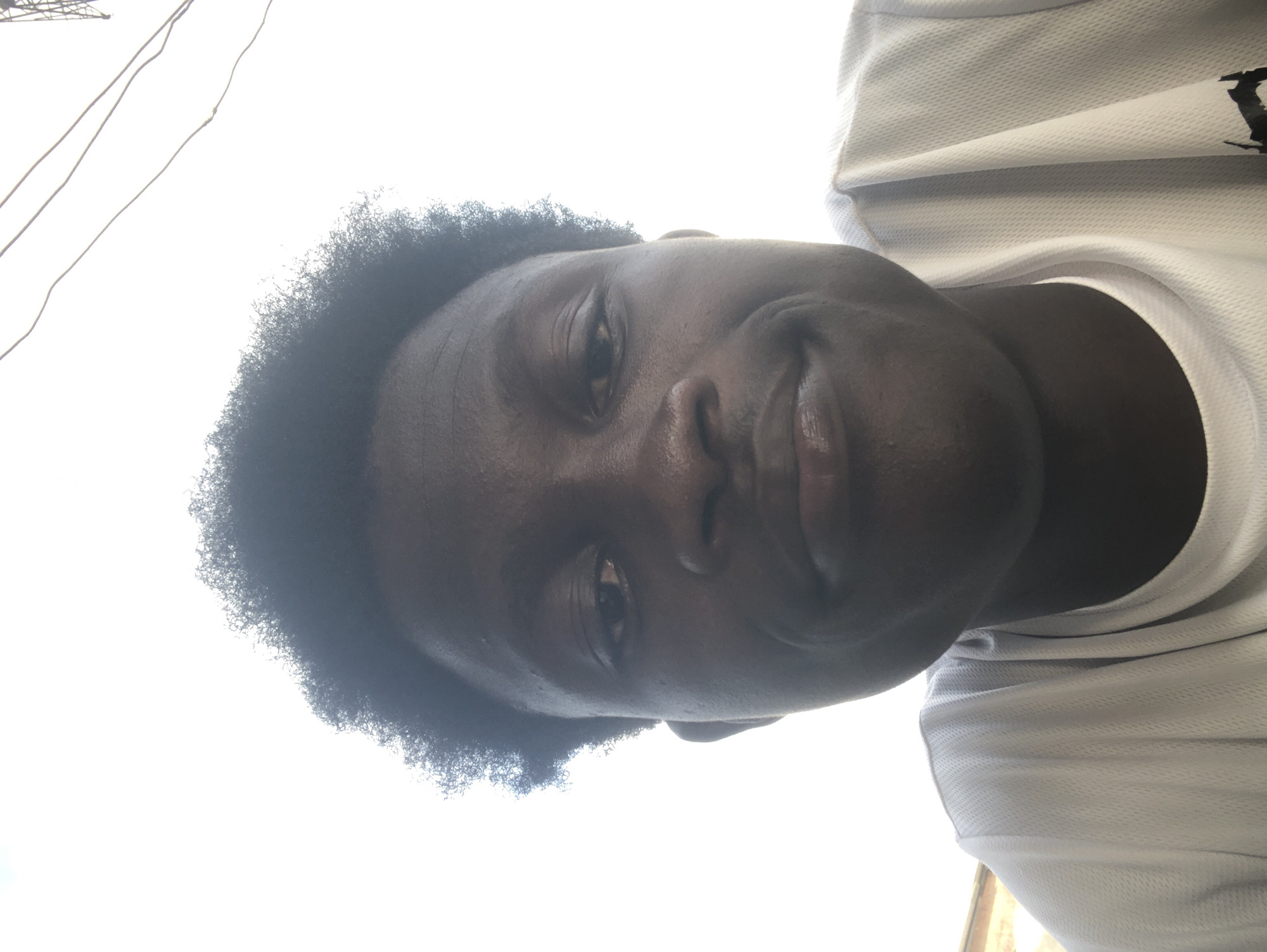 A low-angle, close-up selfie of a young Black male with dark skin and dark, curly hair. He is wearing a white t-shirt. His eyes are open, and he has a slight smile. The background is a bright, overcast sky with some thin wires visible in the upper right quadrant, suggesting an outdoor setting. The image appears to have been taken in Ado, Nigeria.