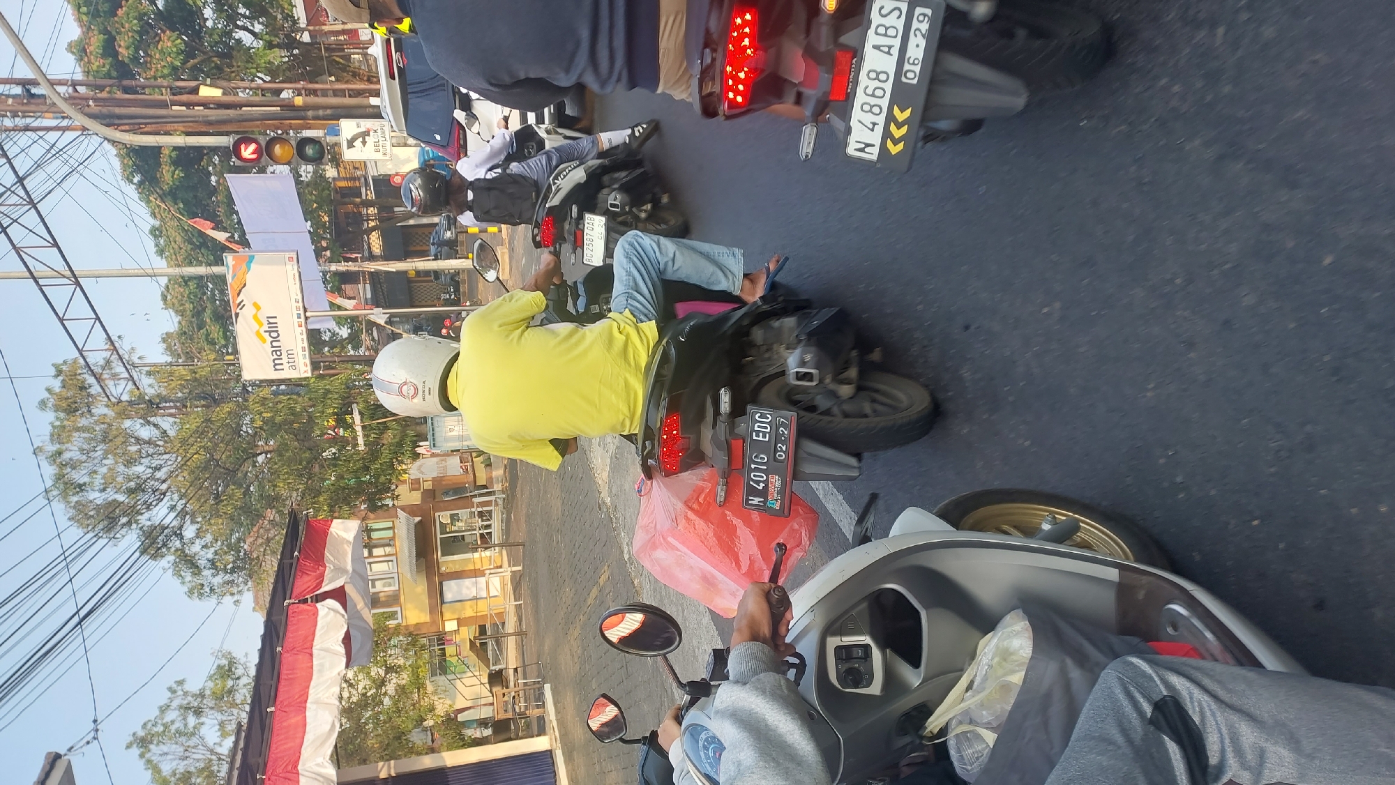 The image depicts a street scene in Lesanpuro, Indonesia, during daylight hours. A traffic jam is evident, with several motorcycles stopped on a paved road. In the foreground, a person on a light-colored scooter is visible from the waist down, wearing grey pants. To their right, a person on a black motorcycle wears a yellow t-shirt and a white helmet, with a large red plastic bag secured to the back of the motorcycle. The license plate on this motorcycle reads "N 4016 EDC". Further back, another person on a black motorcycle with a white helmet is visible, and behind them, a white car is stopped. A traffic light is visible in the background, with the red light illuminated. A sign for "mandiri atm" is also present. Trees and overhead power lines form part of the urban environment.