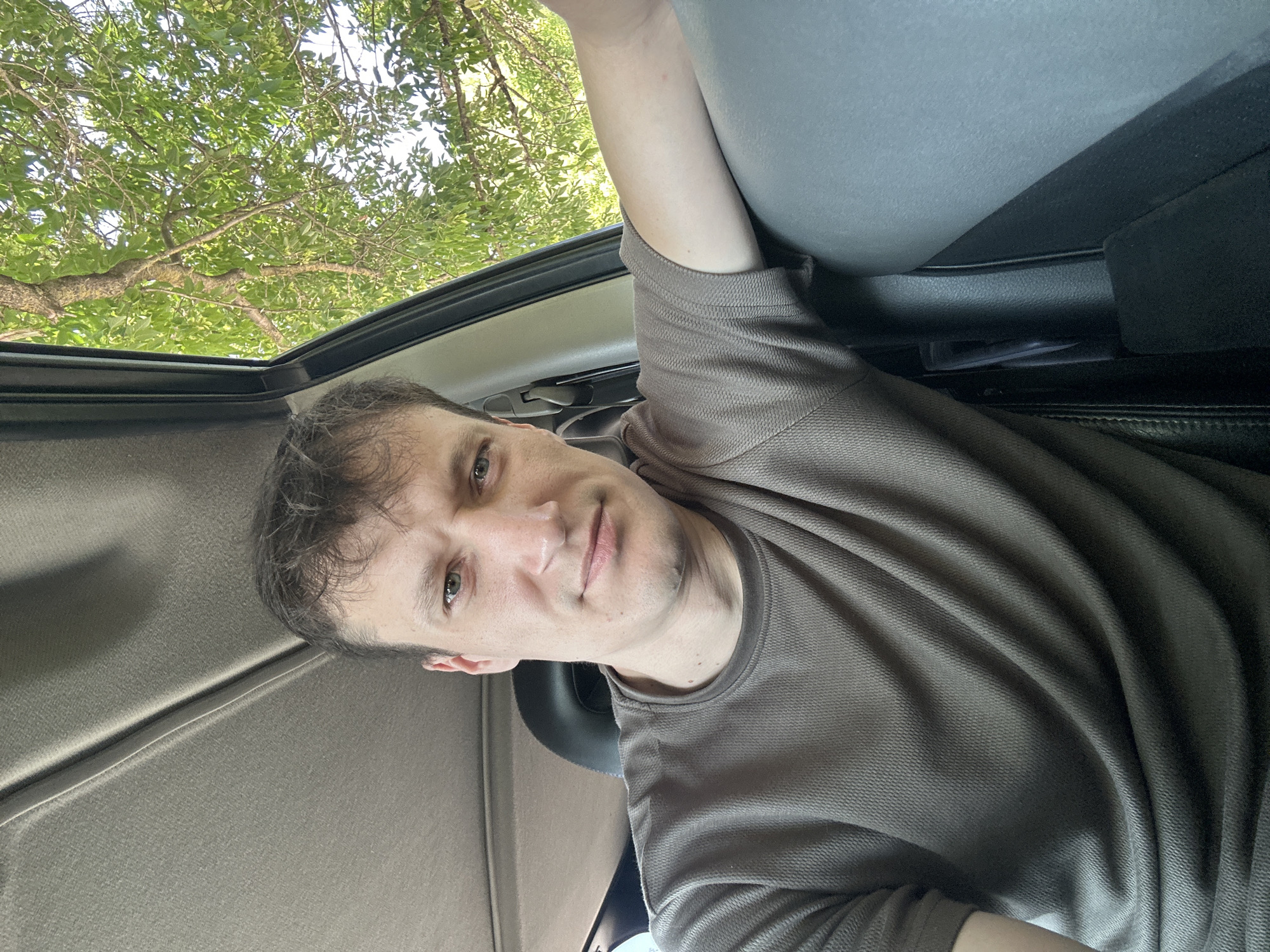 A man is captured in a selfie from inside a vehicle, with his right arm resting on the gray interior. He is wearing a brown t-shirt. Through the open car window, lush green trees and foliage are visible against a bright sky, suggesting an outdoor setting. The background also shows parts of the car's interior, including the headliner and a black headrest. The image does not provide any specific information about the location in the city of Makiv, Ukraine.