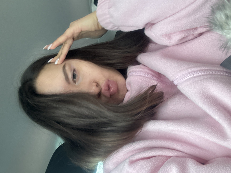 A single female subject with long, dark hair, partially covering the left side of her face, is depicted in what appears to be the interior of a vehicle. Her lips are pursed forward. She is wearing a light pink, textured fleece garment with a visible zipper detail around the collar area. Her right hand is raised, with fingers extended towards her temple; the fingernails are painted white with a pointed shape. The background consists of dark and light gray tones, consistent with a vehicle's headliner and interior trim. No identifying markers for the specific city of Рассудово, Russia, or any other precise geographic location are present within the image.