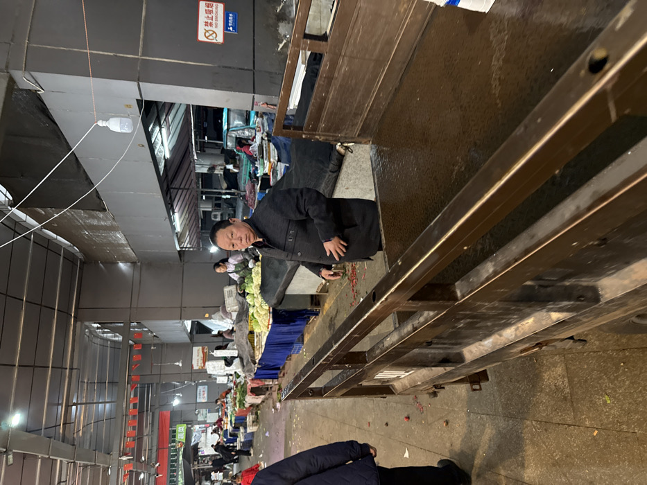 The scene depicts an indoor fresh produce market in Jiangjin, China. In the foreground, the metal-lined bed of a dark brown utility vehicle or truck occupies the right half of the frame, showing a textured, potentially wet or reflective surface.
In the midground, a middle-aged East Asian male with short, dark hair, wearing a dark blue or black collared jacket with a zippered front and two breast pockets, stands behind the truck bed, facing forward with a neutral expression. His hands are clasped near his midsection. To his left, partially obscured by the truck, another individual wearing a dark blue jacket and black pants is visible from the waist down, walking towards the left out of the frame. Red debris, possibly from produce or firecrackers, is scattered on the concrete floor between this individual and the truck.
Behind the central male figure, a market stall is visible, displaying various green leafy vegetables, including what appears to be cabbage. A woman in a light-colored apron stands behind this stall, attending to the produce. Further back, numerous other market stalls are arranged in rows, covered with blue tarps and displaying more produce. Several other individuals are visible in the background, moving between stalls or attending them.
The market ceiling is high, constructed with exposed metal beams and grey structural elements. Multiple white, cylindrical LED light bulbs hang from wires, providing illumination. Red banners with Chinese characters are strung across the market in the upper left background. On a grey pillar to the right of the central man, a red and white sign with Chinese characters and a "no smoking" pictogram is affixed. The overall environment appears functional, with an active market atmosphere.