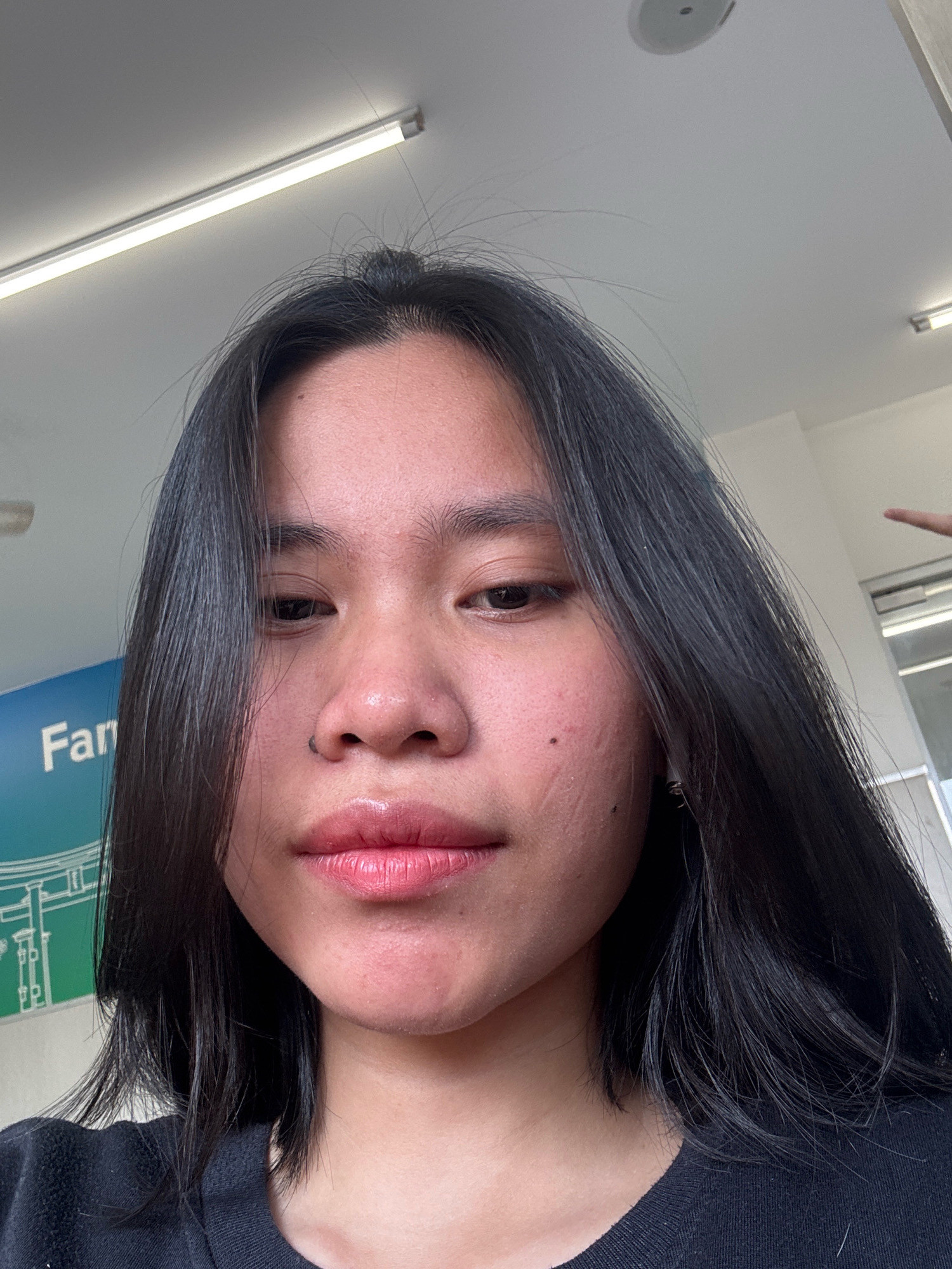 The image is a close-up, selfie-style shot of a young woman with dark hair. She is wearing a black top and has on lip gloss and eyeliner. The background appears to be indoors, with a fluorescent light fixture visible on the ceiling and part of a sign that says "Far..." which could indicate a location in East Jakarta, Indonesia, possibly a business or public facility. The woman is looking directly at the camera. The lighting suggests it is daytime.