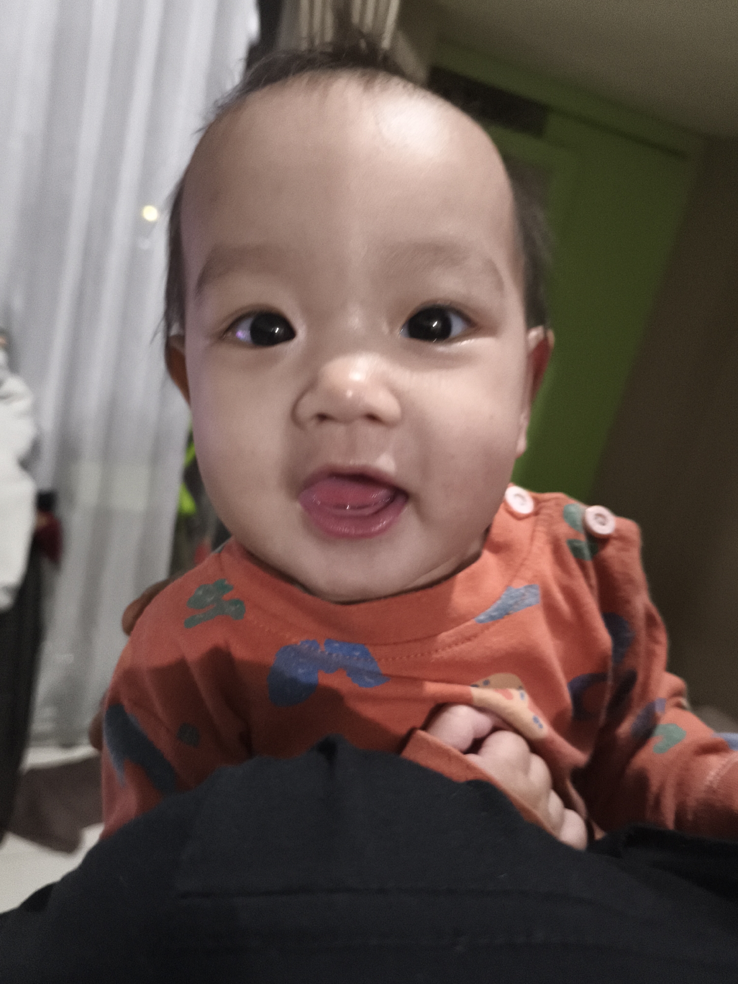 A close-up, slightly blurry photograph captures a baby's face from the chest up. The baby, who appears to be of East Asian descent, has a round face, dark eyes, and is sticking out their tongue. They are wearing an orange long-sleeved shirt with a pattern of blue and green shapes. A person's hand is visible, holding the baby. The background is out of focus and appears to be an indoor setting with draped curtains on the left and a green wall on the right. The lighting suggests it is nighttime or a dimly lit interior. There is no specific indication of the location being in Depok, Indonesia, as the image lacks any recognizable landmarks or context clues.