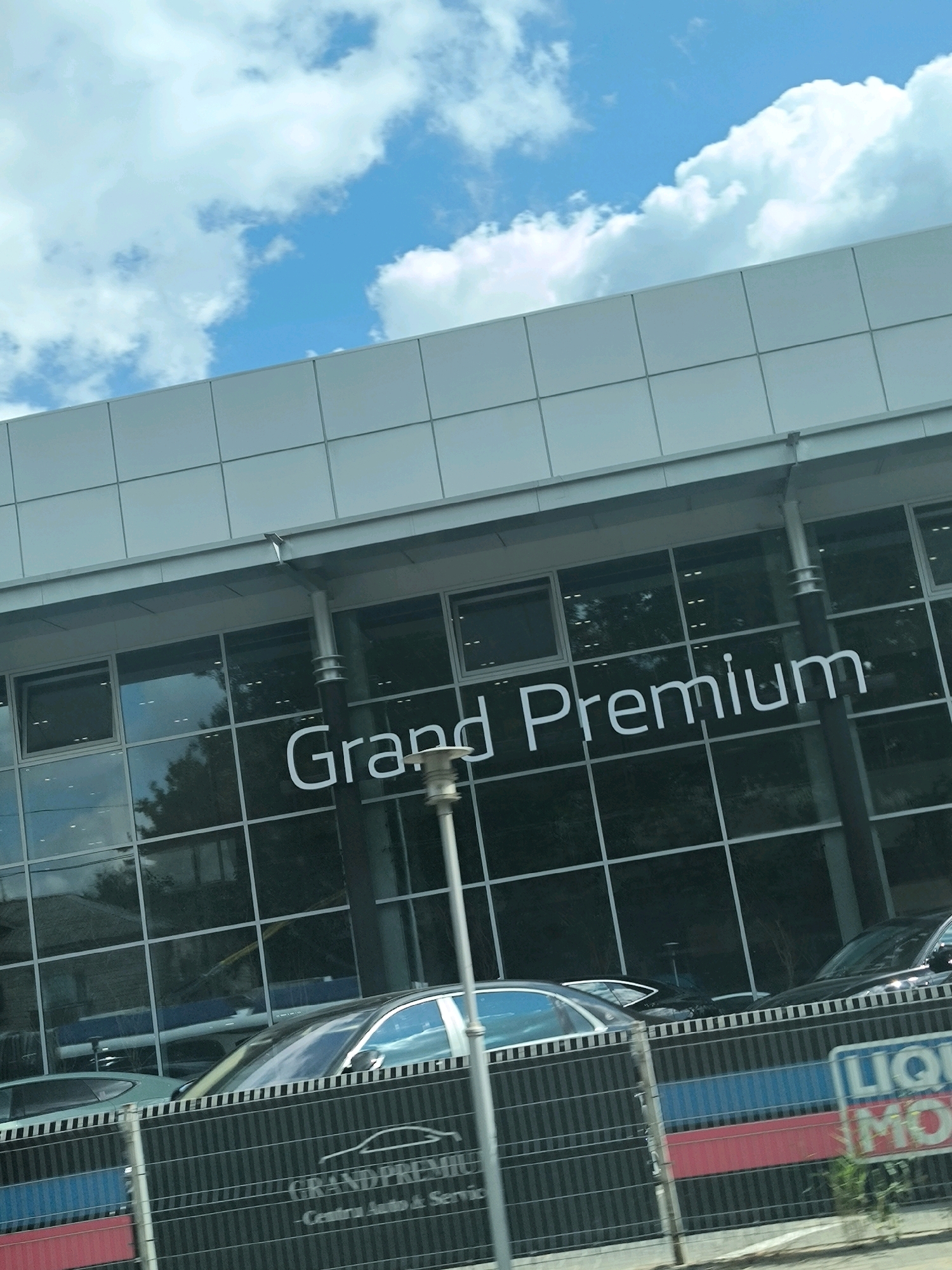 The image captures the facade of a modern building with large glass windows, likely a showroom or dealership, bearing the prominent white lettering "Grand Premium" on its upper section. Above the windows, a section of the building is clad in light-colored rectangular panels, and a clear blue sky with scattered white clouds is visible. In front of the building, a dark fence with a fence top railing is visible, adorned with a striped pattern of black, blue, and red bands. A sign with the logo "LIQUI MOLY" is attached to the fence. Several cars are parked behind the fence and visible through the showroom's windows, including a silver sedan and a black sedan. The overall scene appears to be a bright, sunny day, with the building situated in an urban environment. The presence of the "Grand Premium" signage and the cars suggest it is a car dealership. The "LIQUI MOLY" sign indicates a connection to automotive products or services. Based on external information about "Grand Premium" dealerships, this location is likely in Chișinău, Moldova.