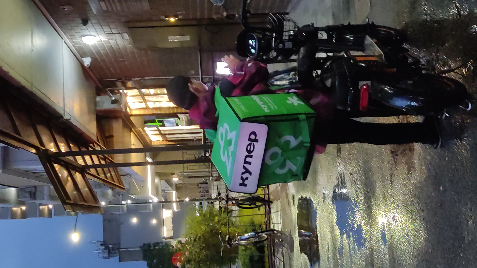 A person wearing a dark jacket and black pants stands next to a black electric scooter, holding a smartphone and looking at it. They are carrying a large, bright green delivery bag on their back, with a white panel on the front featuring a logo and the word "Купер" (Kuper) in black lettering. The bag also has additional logos. The scooter has a basket in front and a visible taillight. The ground is wet and reflects light, indicating recent rain, and puddles are visible. In the background, there's a multi-story building with windows and lights, and what appears to be a covered walkway or awning over a building entrance. The scene suggests an urban environment, likely a street or sidewalk. The image quality is moderate, with a slight blur and low light conditions. The location is identified as Krasnodar, Russia.