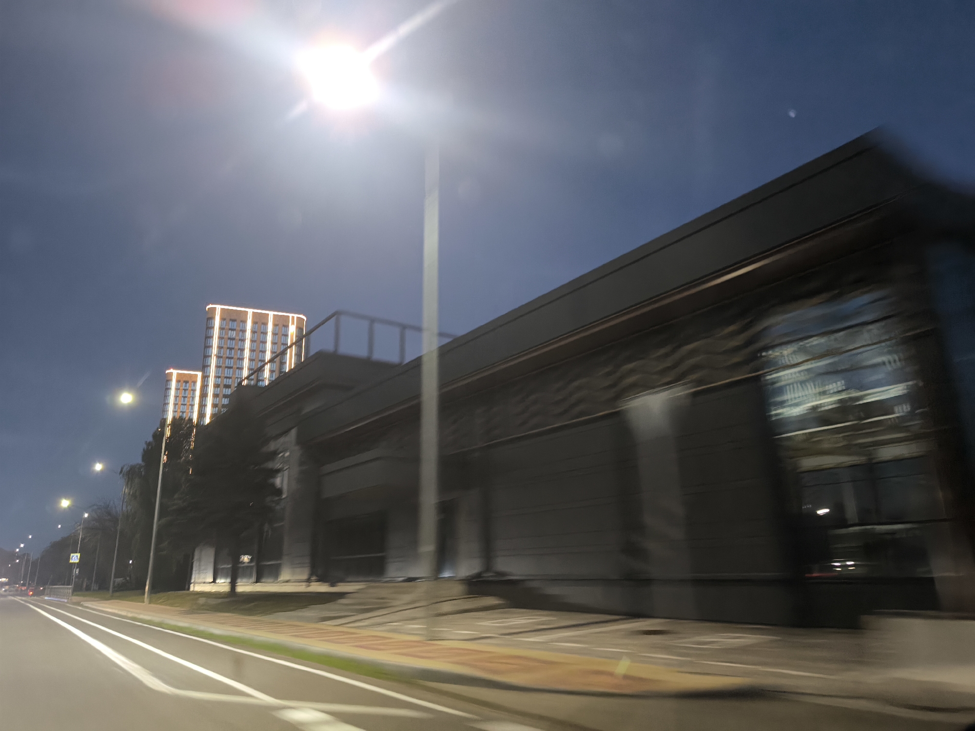The image depicts a street scene at dusk or night in an urban environment. A multi-lane road runs from the left foreground to the background, with white lane markings visible. On the right side of the road is a modern building with a dark, textured facade and large glass windows. The building appears to be a commercial or residential complex, with architectural lighting highlighting its upper floors. Two tall, illuminated residential buildings are visible in the distance to the left, with a streetlamp in between them. Trees are scattered along the roadside. The sky is dark blue with some light haze, and a bright light source, possibly a streetlamp or a very bright light in the sky, is prominent in the upper left quadrant, causing lens flare. The image appears to be taken from a moving vehicle, indicated by motion blur.