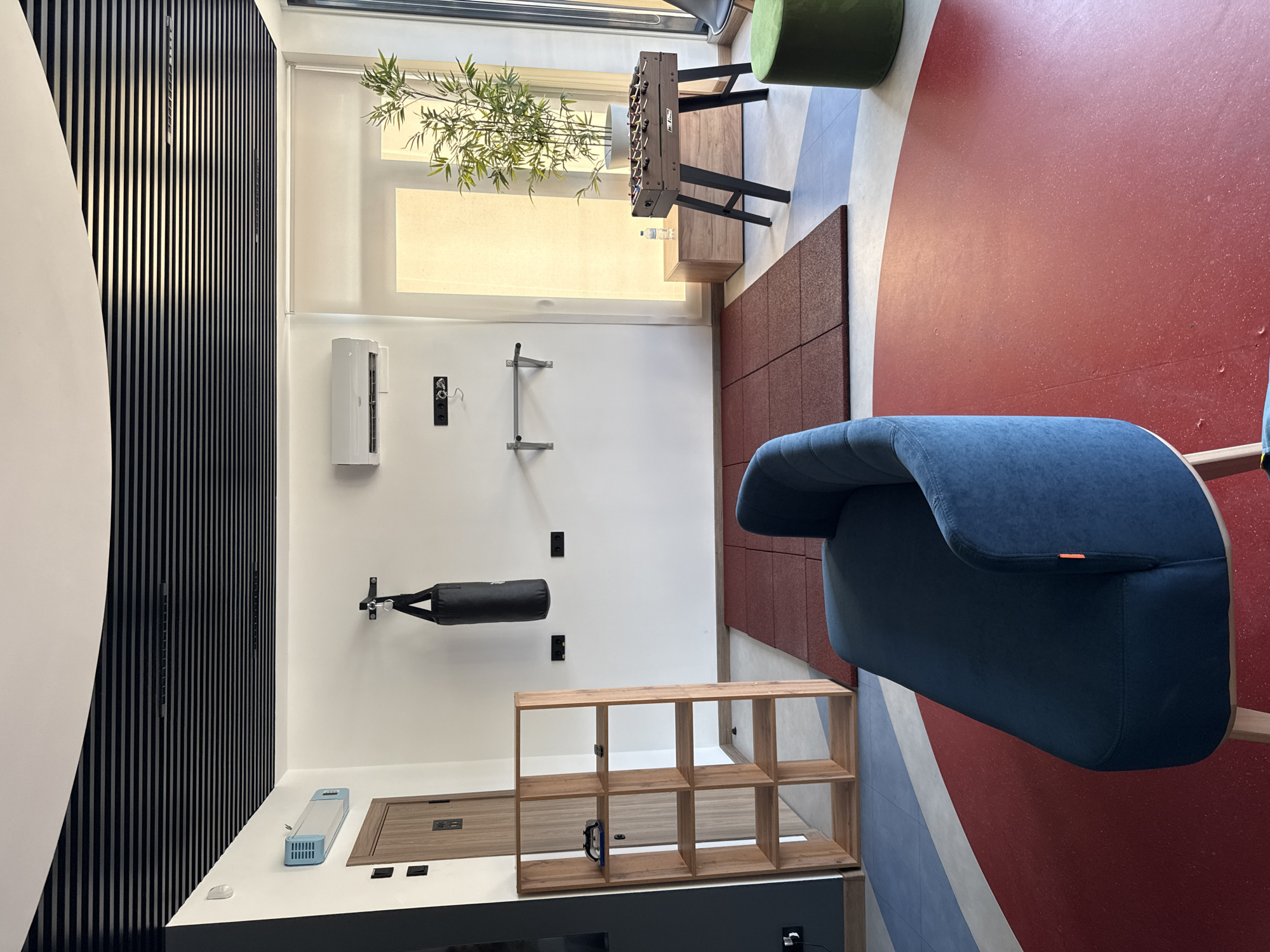 This image depicts an indoor gym or recreational space. In the foreground, a dark blue upholstered sofa is positioned on a red and white circular floor pattern. Behind the sofa, a wooden shelving unit with multiple compartments stands against a white wall. A black punching bag hangs from the wall above a set of red floor mats. To the right, a foosball table is set up next to a wooden cabinet, and a potted bamboo plant is visible behind it. A large window with a roller blind is to the right of the foosball table. The ceiling features a modern design with black horizontal slats. An air conditioning unit is mounted on the white wall. The scene appears to be in Sochi, Russia, based on the context provided. There are no people visible in the image.