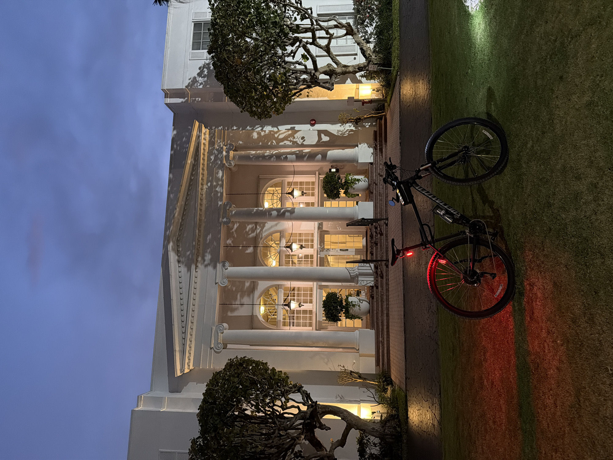 A black Specialized bicycle is parked on a lawn in front of a white neoclassical building at dusk. The building has a grand entrance with large columns, arched windows, and hanging lanterns. The bicycle has a red taillight illuminated, casting a red glow on the grass. A white light from another source illuminates a patch of the lawn to the right of the bicycle. The sky is a deep blue with some clouds. The scene appears to be in Bal Harbour Village, United States of America, likely a hotel or similar establishment given the architecture. There are no people visible in the image.