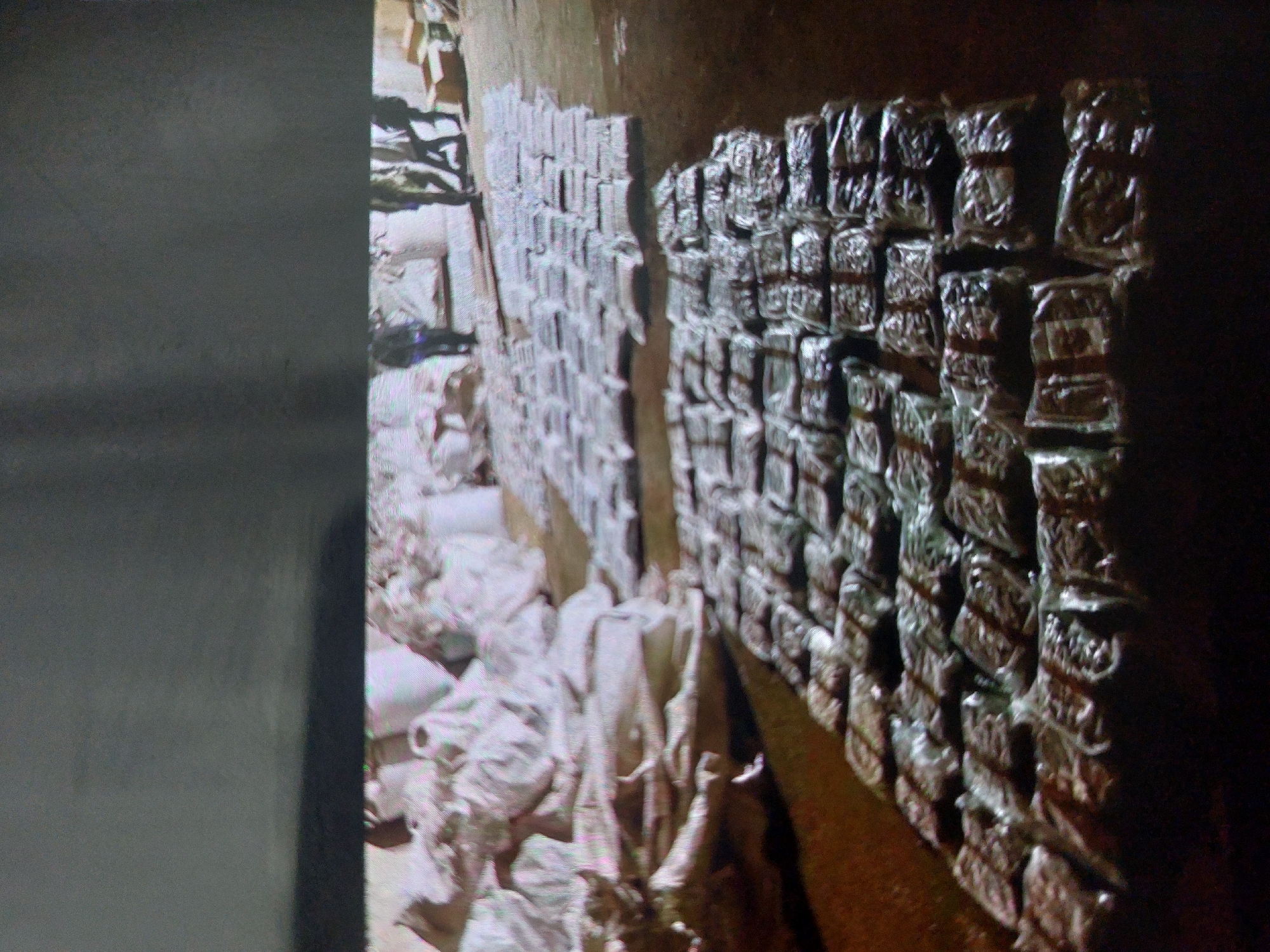 The image shows a dark, possibly indoor location in the city of Tsarevo, Bulgaria, likely a storage facility or warehouse. The scene is filled with stacked objects. On the right, there are stacks of packaged items wrapped in silver material, arranged in neat rows against a wall. These appear to be brick-shaped packages secured with bands. To the left, there's a large pile of white sacks or bags, which appear to be irregularly shaped and possibly filled with soft material. In the far left, there is a grey wall. The lighting is uneven, with some areas in shadow and others more illuminated. There is no evidence of people present in the image. The environment appears to be enclosed and confined.