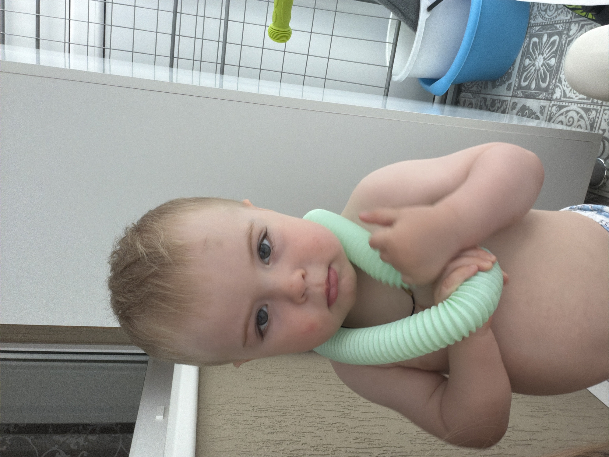 A baby with light blonde hair and blue eyes is holding a green corrugated plastic tube around its neck, possibly in Votkinsk, Russia. The baby is partially unclothed. The background includes a window, a white surface, and a tiled area with a blue bowl.