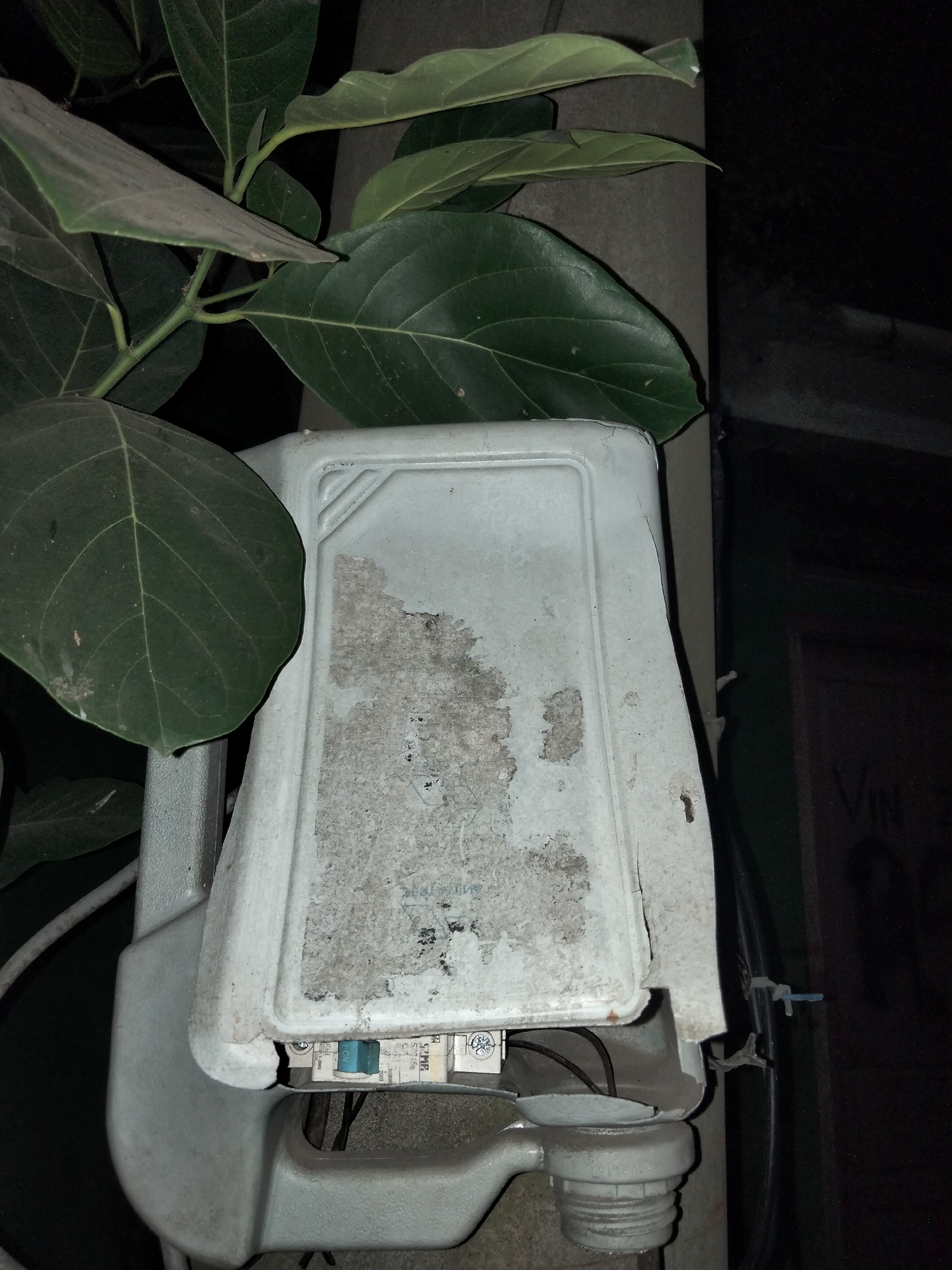 In Cariu, Indonesia, an outdoor electrical junction box, weathered and covered in dust and grime, is mounted on a pole. The box is partially obscured by the large, dark green leaves of a tree. Visible within the open portion of the box are several electrical breakers, one of which is a small blue switch labeled "ON". The background is dark, suggesting nighttime, with a hint of a building or wall visible on the right side, featuring some faint lettering. The scene appears to be a typical urban or semi-urban environment in Indonesia.