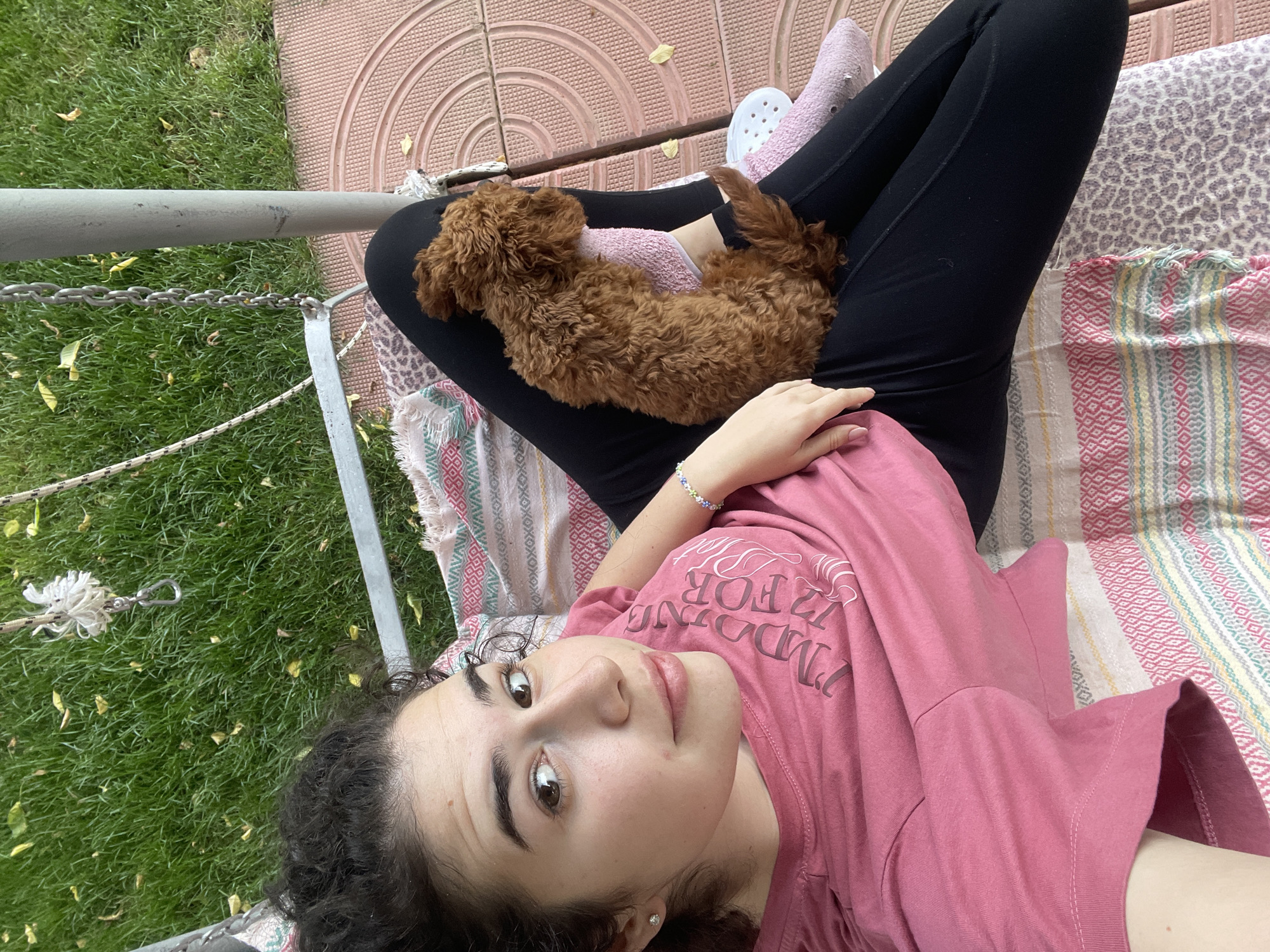 A medium, eye-level shot shows a young woman lying in a hammock with a small brown poodle resting on her lap. The location appears to be outdoors, in a backyard or garden, evidenced by the presence of grass and vegetation in the background.
The woman is wearing black leggings and a pink t-shirt. Her hair is dark and curly. She is looking directly at the camera with a neutral expression. A bracelet is visible on her left wrist.
The poodle is lying comfortably on the woman's legs, curled up.
The hammock is patterned with stripes and geometric designs. Part of the hammock frame is visible on the left side of the image, made of metal.
Based on the architecture and vegetation, there's no specific landmark in the image that allows pinpointing the exact location in Sofia, Bulgaria.