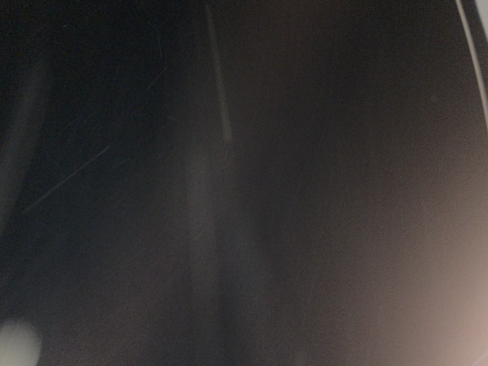 A dark, close-up photograph displays a grainy, dark grey surface. The image is rich in digital noise, suggesting it was captured in low light conditions or with a high ISO setting. Several thin, linear scratches are visible on the surface, primarily in the upper-left and lower-left portions, indicating wear or damage.
Various light reflections and glows are present across the dark background. A subtle, vertical streak of brighter light extends downwards from the top-center. In the bottom-left corner, a larger, diffuse blob of bright, out-of-focus light is visible. A distinct, brighter vertical line runs along the far right edge of the frame, appearing as a sharp reflection. Additionally, a soft, warm-toned glow emanates from the bottom-right corner. There are no discernible objects, people, or activities taking place. No text is visible. The image provides no specific cues about the time of day, weather, or explicit location within Karamsar, Pakistan.