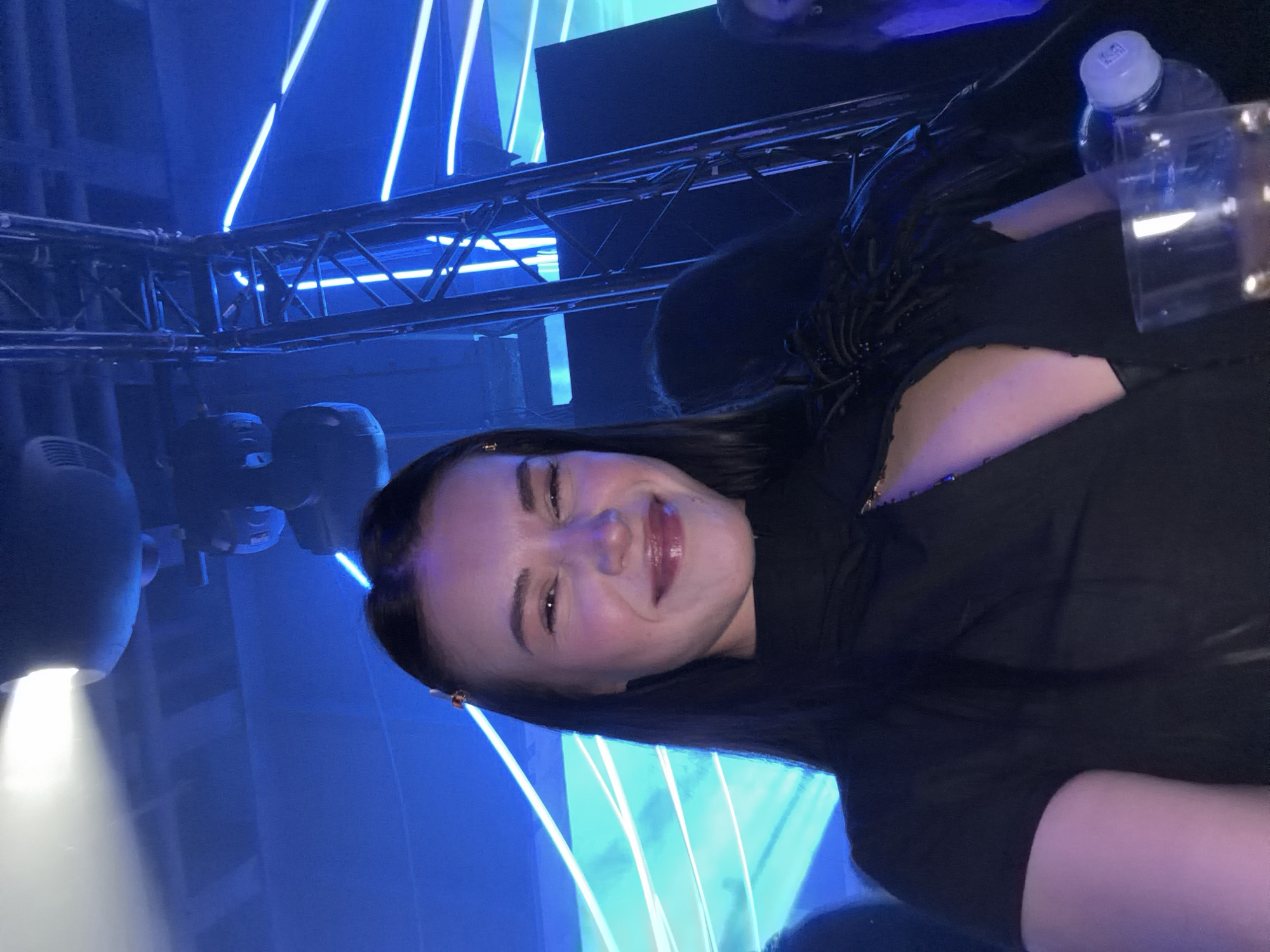 A photograph taken indoors depicts a woman with dark hair and fair skin wearing a dark top. She is smiling. The background includes a lighting rig with blue neon lights. A water bottle and a clear plastic cup are visible in the upper right corner. The photograph appears to be taken at an angle in a nightclub or event space setting. It is not possible to ascertain the location of the scene in Moscow, Russia from the image.