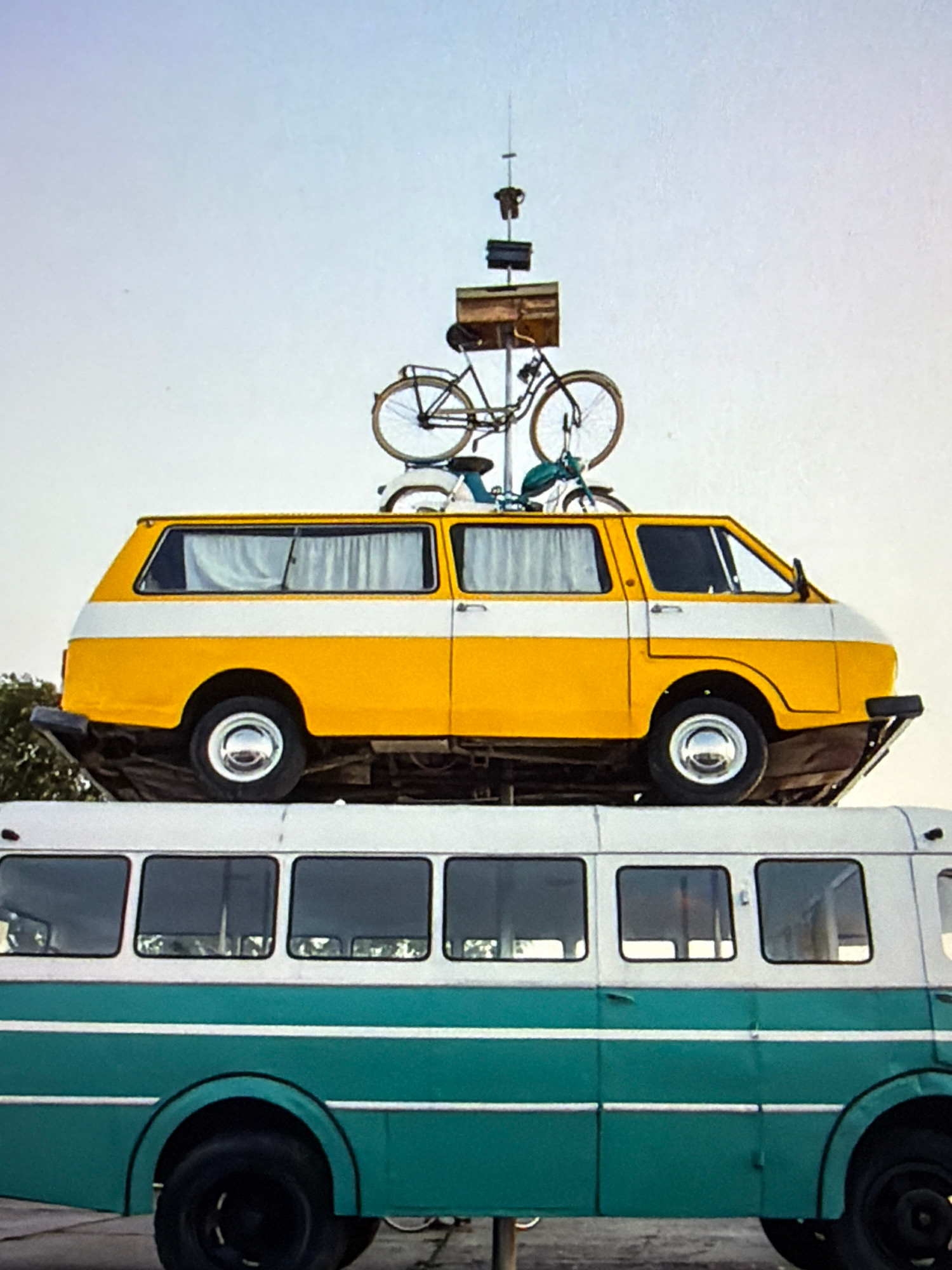 In Kafr Yasif, Israel, an unusual art installation features a yellow and white station wagon balanced on top of a teal and white bus. The station wagon is further adorned with two bicycles mounted on its roof, one of which is resting on top of the other. Above the vehicles, a thin pole extends upwards, supporting a small structure that appears to be made of wood and metal, with what looks like a dark shirt hanging from it. The background consists of a pale, clear sky. The overall scene is static, with no visible human presence or activity.