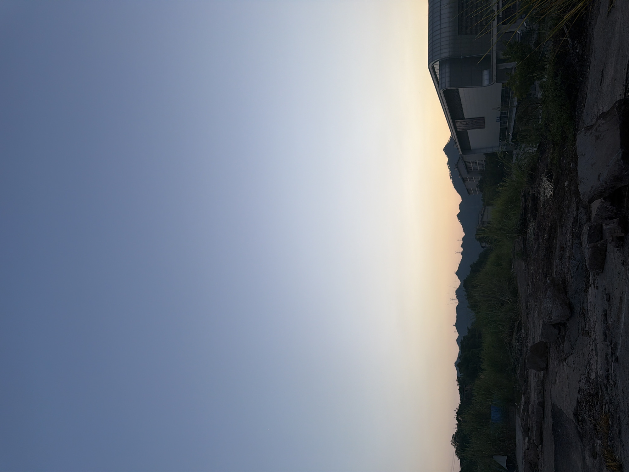 This is a landscape photograph taken during the twilight hours in Jiangjin District, Chongqing, China. The sky transitions from a deep blue at the top to a soft orange and yellow hue near the horizon, indicating either sunrise or sunset. In the foreground, there is an unpaved, rocky area with scattered debris, suggesting a construction site or an undeveloped patch of land. Tall, green grass grows in clumps throughout this area.
Dominating the right side of the mid-ground is a modern building with a distinctive curved, metallic roof and large glass windows. To the left of this building, and behind the dense grass, are silhouetted trees and distant hills or mountains. Some power lines and transmission towers are visible against the fainter sky above the mountains. The overall scene depicts an urban fringe environment with elements of both nature and development under a serene, atmospheric sky. There are no people visible in the image.