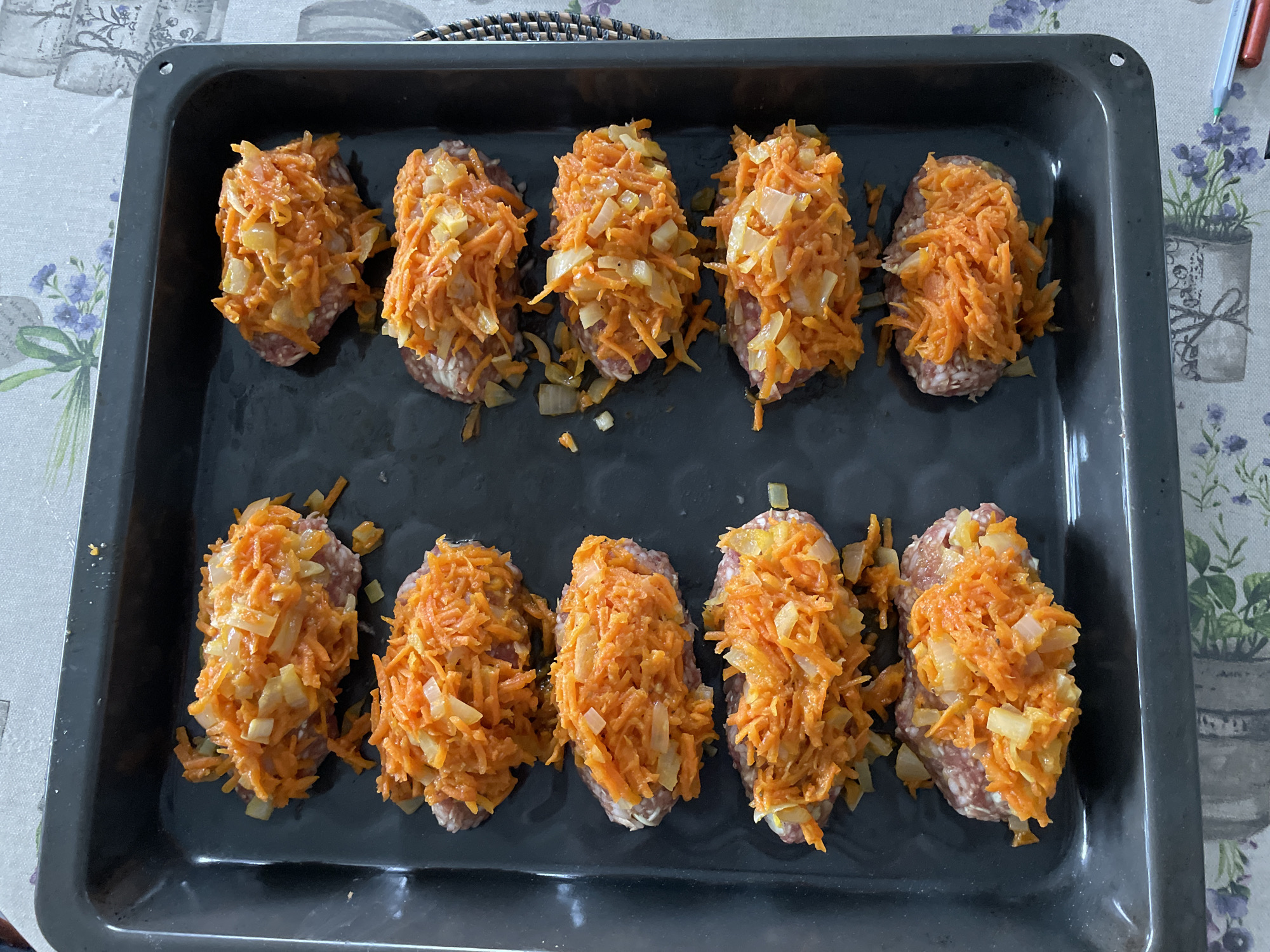 A top-down shot shows a black baking tray filled with ten oblong-shaped portions of meat topped with shredded carrots and diced onions. The tray sits on a table with a patterned tablecloth. The scene is in Лесной Пейзаж Кп (шарапово, Russia.
