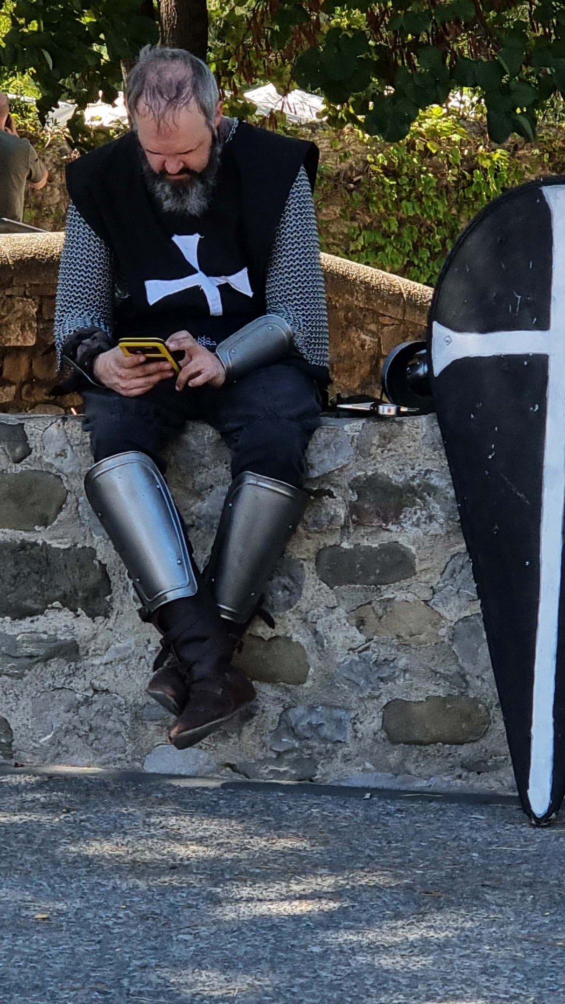 A man dressed in medieval armor, including chain mail, arm guards, and leg guards, sits on a stone wall in Besalú, Spain. He has a grey beard and is looking down at a yellow smartphone he is holding. A black shield with a white cross is propped against the wall to his right. The background shows greenery and tents, suggesting an outdoor event or festival. The wall is made of rough-hewn stones.