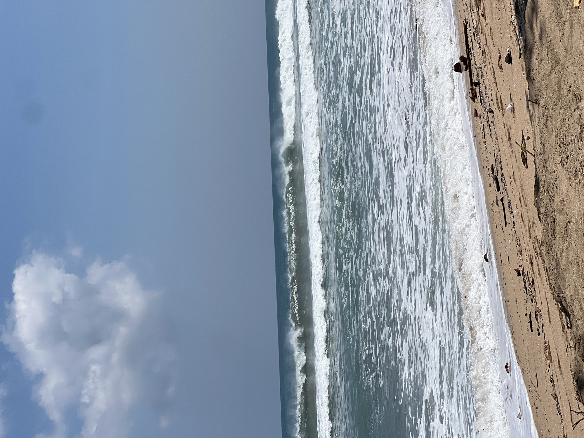 The image depicts a beach scene in Kuta, Indonesia. The foreground is a sandy beach with scattered debris, including sticks, plastic fragments, and what appear to be small pieces of organic matter. A gentle wave is washing ashore, leaving a foamy white line on the wet sand. In the midground, ocean waves are rolling in, with some breaking and forming whitecaps. The water is a greenish-blue color. The background shows a clear blue sky with a single fluffy white cloud on the left side. The horizon line is visible, separating the sea from the sky. The overall impression is a natural coastal environment with signs of human impact due to the debris on the beach.