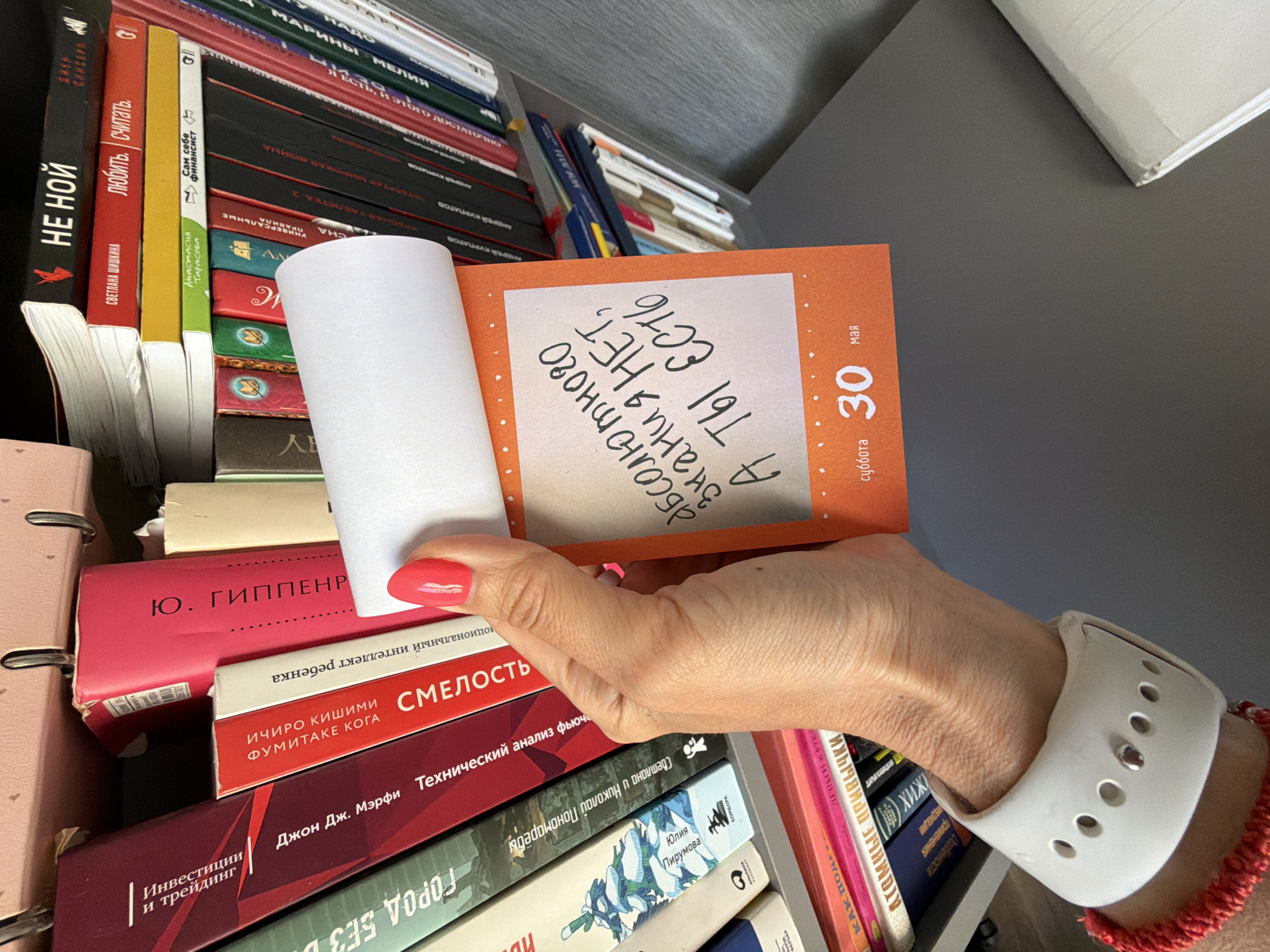 A close-up shot shows a person's hand holding an orange calendar page with the text "There is no absolute knowledge, you always need to learn." The calendar indicates the date as "Saturday, May 30." The hand has a white smartwatch band and a red bracelet. In the background, there is a bookshelf filled with various books, including titles such as "Don't whine" by Svetlana Shishkina, "Investments and trading", "Atomic habits", and "City without boundaries". The books are in Russian and English. The scene appears to be indoors, possibly in a home or office setting. Birobidzhan, Russia is not visible in the image.