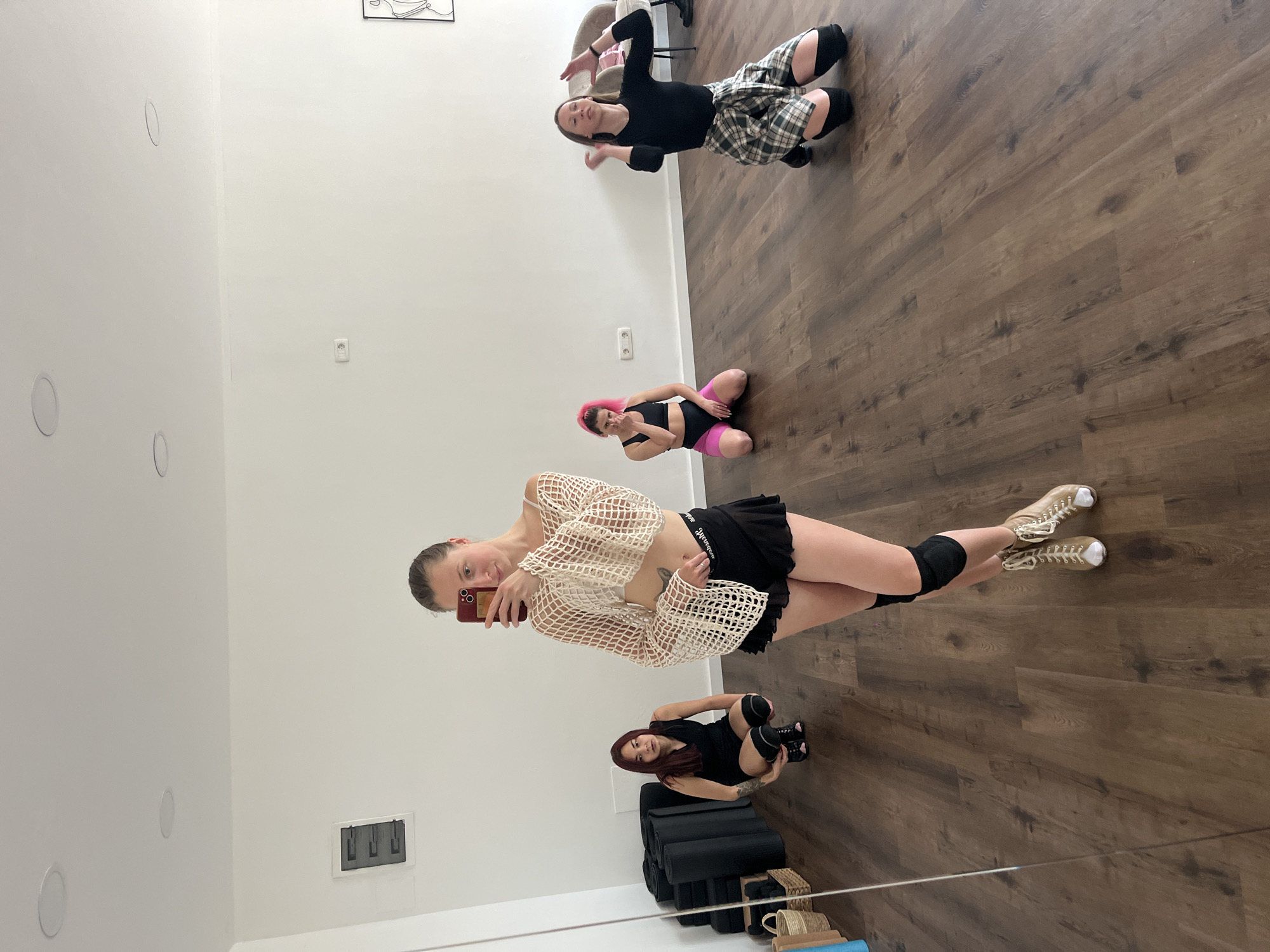 The image depicts an indoor dance or fitness studio located in Budva, Montenegro. Four adult females are present in various poses on a dark wood-patterned floor.
In the foreground, a female with light-brown hair pulled back stands, taking a mirror selfie with a red smartphone. She wears a beige net-like long-sleeved crop top over a white bra, black ruffled shorts with a visible "Calvin Klein" waistband, black knee pads, and light-brown high-top lace-up shoes. A tattoo is visible on her left upper thigh/hip area.
To her immediate left, reflected in the full-length mirror, a female with long, dark red hair is in a low squatting position. She wears a black sleeveless top, black shorts, black knee-high socks, and black dance shoes.
In the mid-ground, slightly to the left of center, a female with bright pink hair kneels on the floor. She is dressed in a black sports bra and pink and black athletic shorts, with her right hand near her chin.
Further to the right, another female kneels on the floor, facing forward with her arms slightly raised in a dynamic pose. She wears a long-sleeved black top, a plaid shirt tied around her waist, and black knee pads.
The studio features plain white walls, a white ceiling with multiple recessed circular light fixtures, and the dark wood-patterned floor. On the left, stacked black rolled mats, a woven basket, and a blue rolled mat are visible against the wall. An electrical panel with a black cover is mounted on the wall near the mirror, and a white electrical outlet is visible on the back wall. In the far right background, a minimalist black wireframe art piece hangs on the wall, and part of a light-colored armchair is visible.