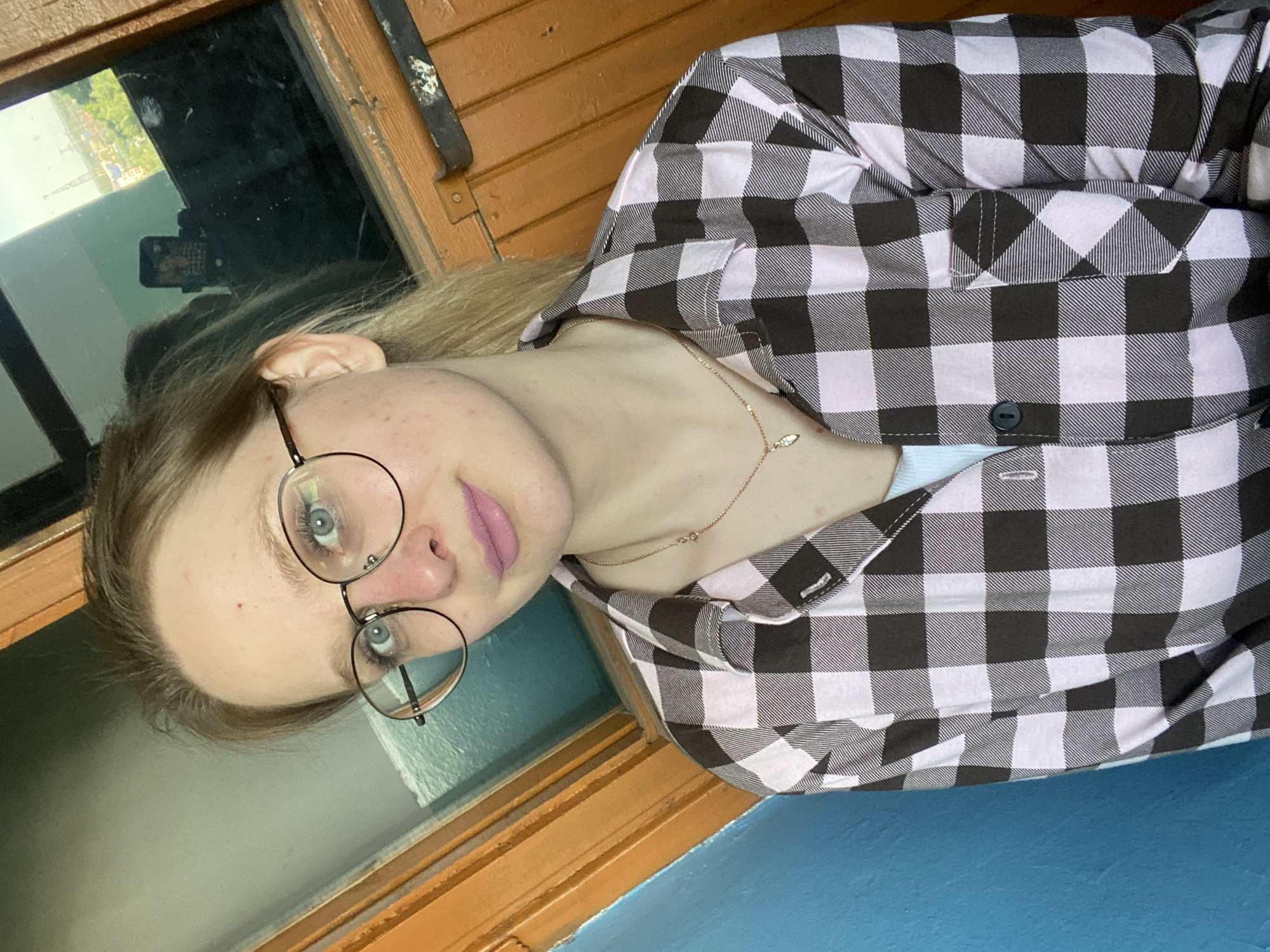 A young, fair-skinned woman wearing round eyeglasses and a black and white checkered shirt is pictured. She is also wearing a delicate gold necklace. The background shows a window with a reflection of what appears to be a city. The window frame and the surface below are painted in orange and blue, respectively. The city appears to be located in Unknown, Unknown.