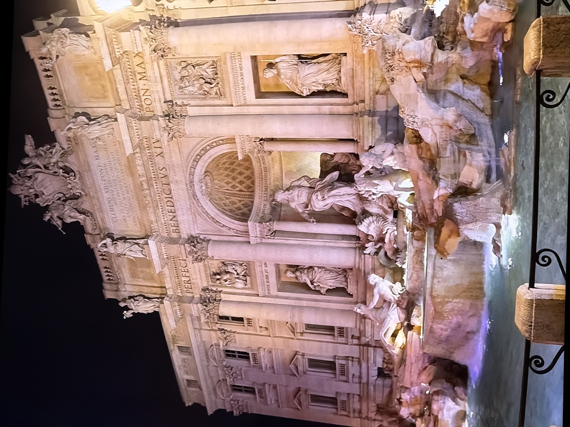 The image depicts the Trevi Fountain at night, illuminated by artificial lighting that casts a warm glow on the Baroque architecture. The fountain is a massive structure with numerous sculptures of mythological figures, predominantly rendered in white marble, set against a grand facade. Water cascades over rocky outcroppings and into a pool in the foreground. The lighting creates vibrant colors in the water, with areas of purple and turquoise. The scene is framed by a dark, shadowy sky above and a black metal railing with decorative scrollwork in the lower portion of the image. The overall impression is one of grandeur and dramatic illumination.