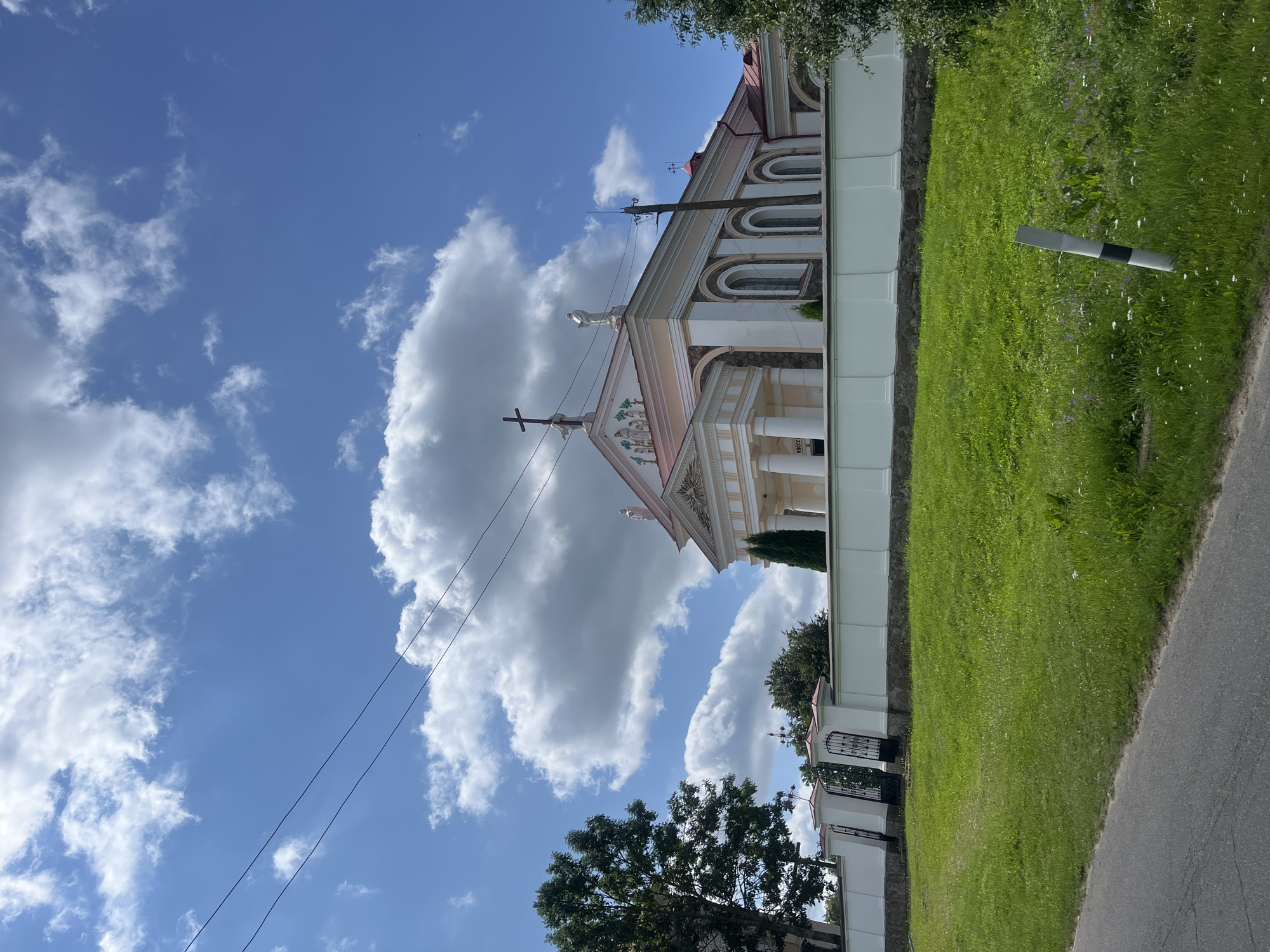 The image shows a large, white church with a pale pink trim, located in the city of Zhaludok, Belarus. The church features classical architectural elements, including a pediment, columns, and arched windows. A cross is visible on the roof, as are statues and decorative friezes. The church is surrounded by a white wall topped with decorative elements, and ornate black gates are visible in the wall. A lush green lawn with scattered wildflowers is in the foreground, with a paved road to the left. The sky is blue with fluffy white clouds, and power lines are visible stretching across the sky. A tall green tree stands to the left of the church. The overall scene depicts a sunny day in a peaceful, historical setting.