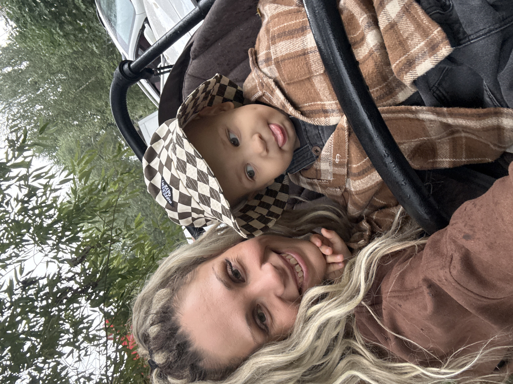 In Rusanovo, Russia, a woman with blonde, wavy hair and a brown top is captured in a selfie with a young child in a stroller. The woman is smiling at the camera, resting her chin on her hand, while the child, wearing a plaid hat and a plaid shirt over overalls, sticks out its tongue. The stroller is dark brown, and a black bar runs across the foreground. In the background, green trees and the side of a white car are visible, suggesting an outdoor setting.