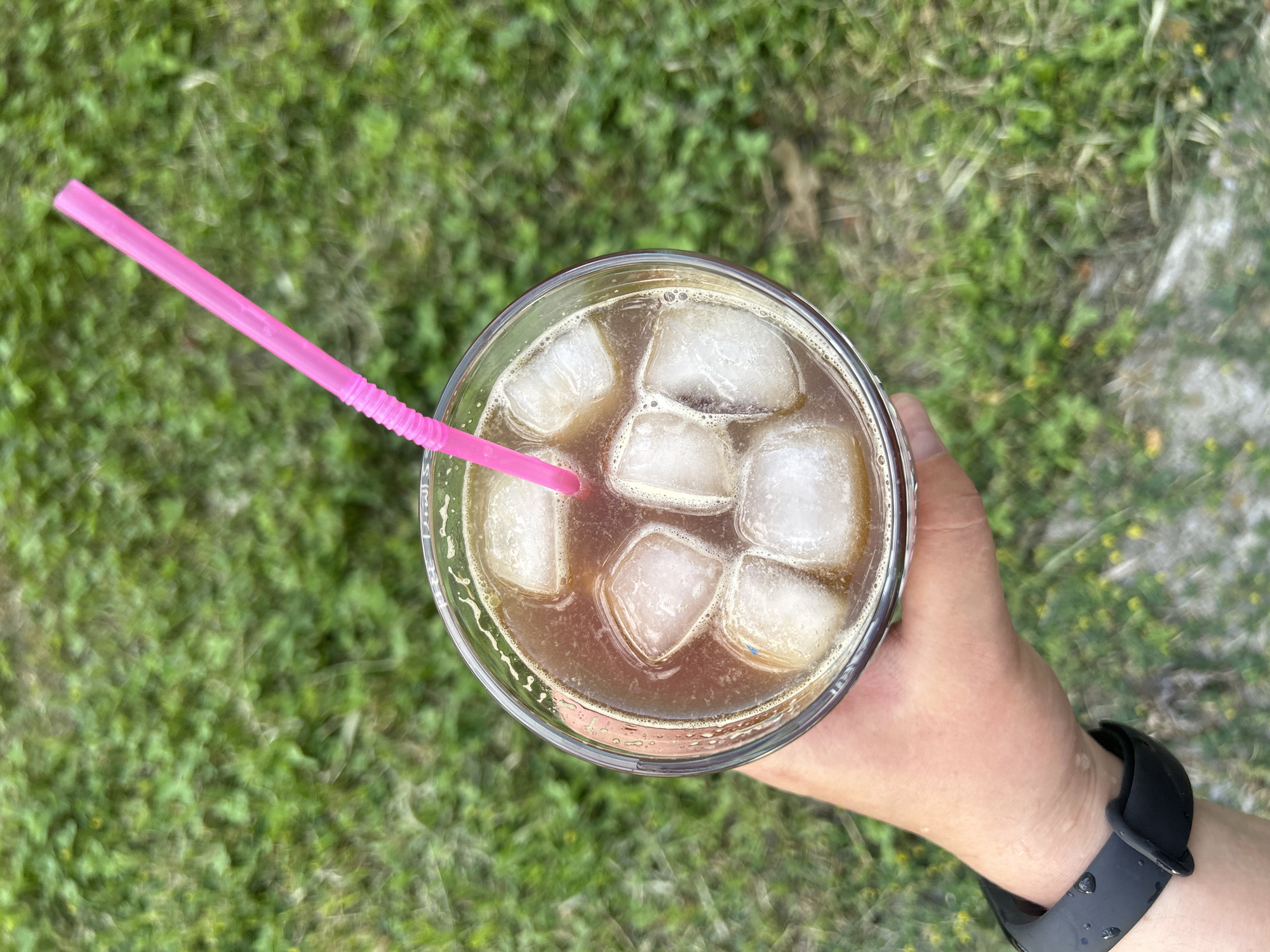 A hand wearing a black fitness tracker holds a clear plastic cup filled with iced coffee and a pink straw. The cup is positioned over a background of blurred green grass, suggestive of an outdoor setting. The scene is viewed from above.