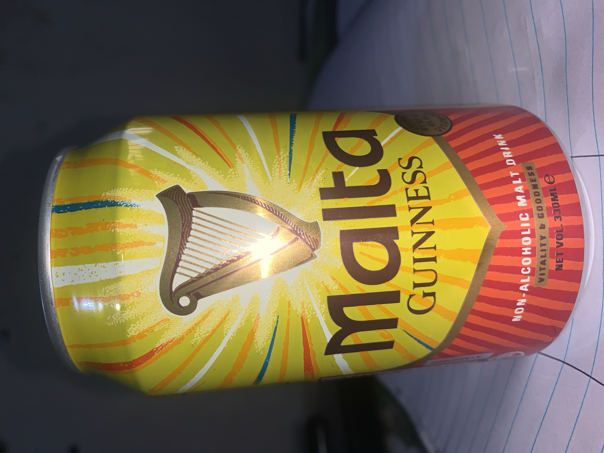 A can of Malta Guinness sits on a lined surface. The can has a yellow background with a golden harp emblem and the text "Malta GUINNESS" in large, brown letters. Red and orange stripes are at the top of the can, along with the text "NON-ALCOHOLIC MALT DRINK VITALITY & GOODNESS NET VOL. 330ML e". The city or specific location of New Stadium, Nigeria, is not represented in the image.
