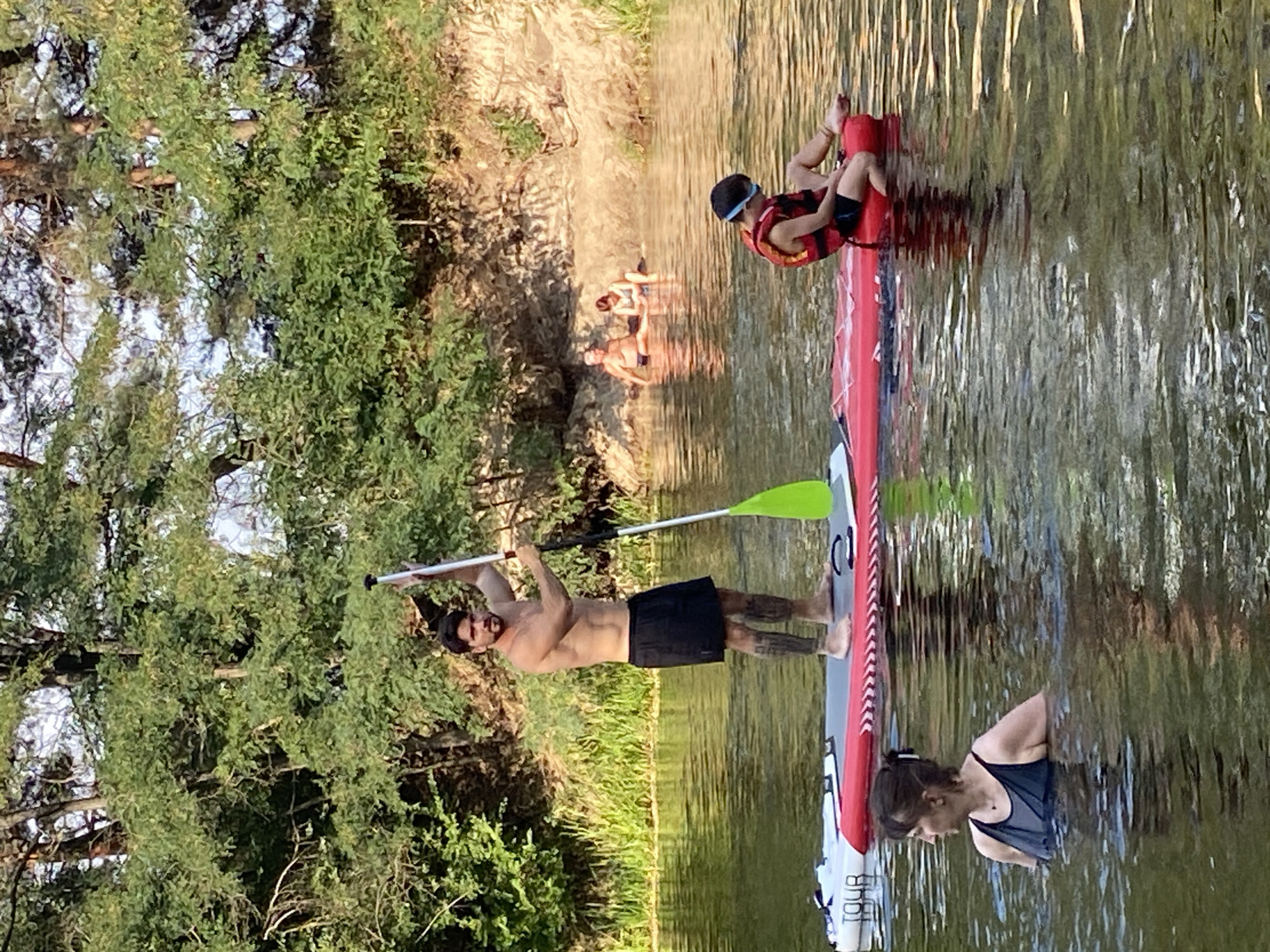 In the city of Semianivka, Ukraine, on a sun-drenched day, a group of people enjoys recreational activities on a calm body of water, likely a river, bordered by lush green trees and a sandy embankment. In the foreground, a woman with dark hair pulled back in a bun, wearing a dark swimsuit, stands waist-deep in the water, looking to her left. To her right, a young person, wearing a life vest and goggles, sits on the edge of a red inflatable raft. Further back, a man with a muscular physique, wearing black shorts, stands on a white and red paddleboard, holding a paddle with a bright green blade. His left arm is raised, and he appears to be looking towards the left side of the frame. In the background, two individuals, a man and a woman, are seated on the sandy bank, partially submerged in the water. The water reflects the sunlight, creating a shimmering effect, and the overall scene conveys a peaceful, leisurely atmosphere.