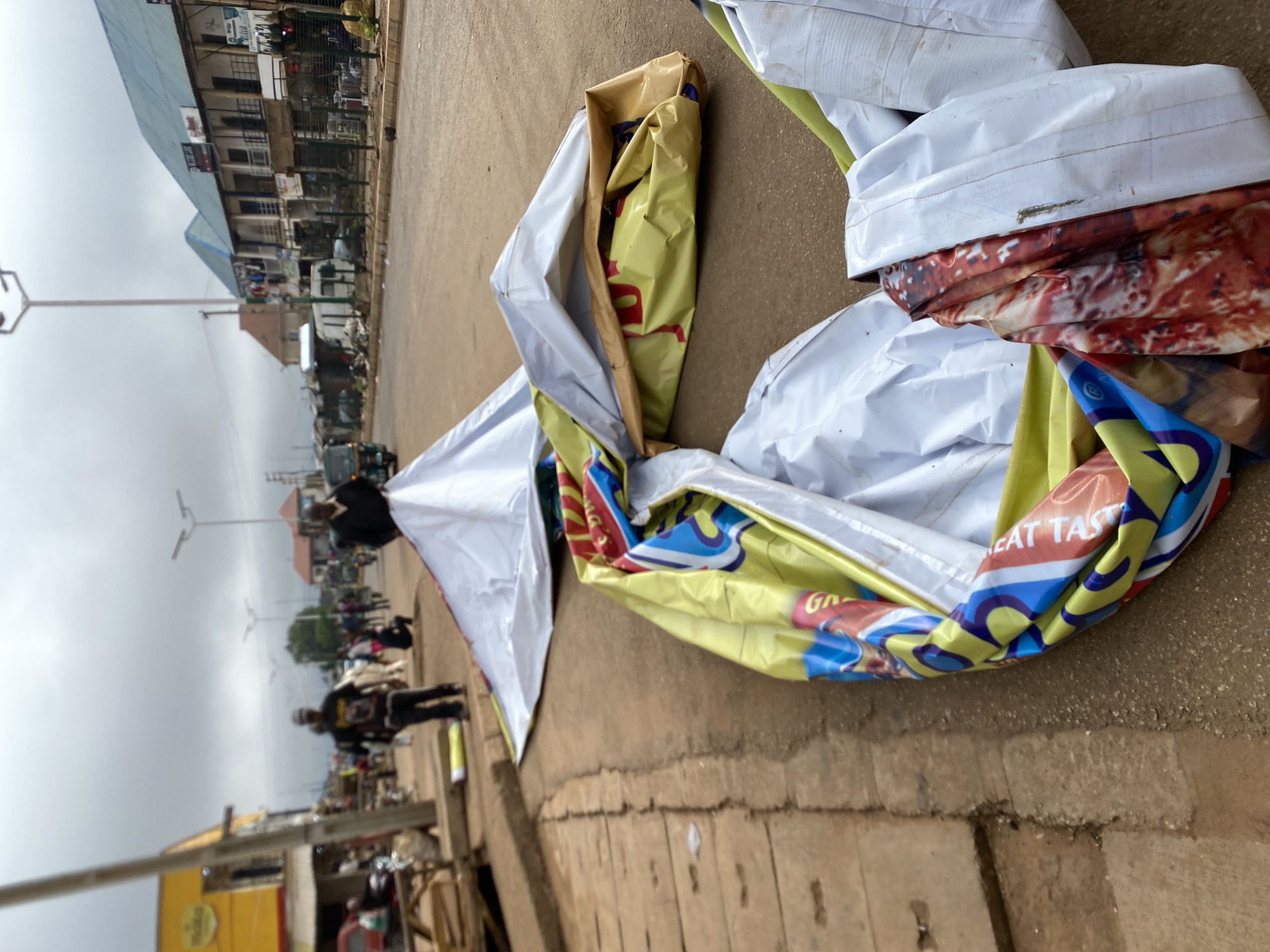 The image shows a discarded vinyl banner lying on a street in Bukuru, Nigeria. The banner is predominantly white, yellow, and blue, with portions showing food imagery and text. It appears crumpled and partially folded. In the background, the street is lined with buildings, some with multiple stories. There are people walking on the street and parked vehicles. Street lights are visible. The sky is overcast.