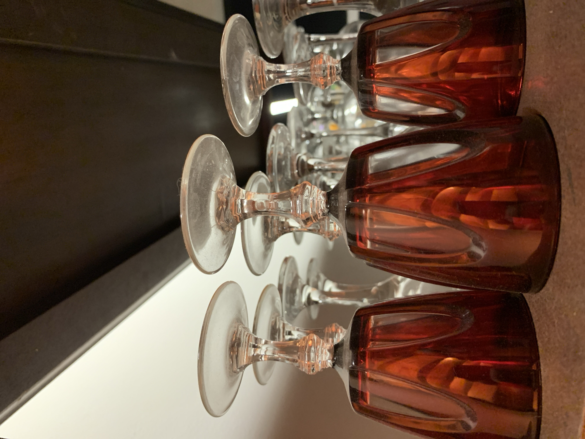 The image presents a close-up, low-angle perspective of numerous stemmed glasses stored inverted on a textured, brownish-gray surface. In the foreground, three prominent goblets feature deep reddish-amber, faceted bowls and clear, intricately detailed stems with circular bases facing upwards. These amber glasses are inverted, resting on their rims. Behind them, several more clear glass, stemmed goblets are visible, also inverted, extending into the midground and background, creating a sense of depth. The clear glass elements show minor dust accumulation on the upward-facing bases.
The background consists of a bright, off-white vertical surface to the upper left, potentially a wall or interior cabinet. To the upper right and directly above the glasses, a dark brown, possibly wooden, horizontal surface is visible, likely an overhead shelf or the interior ceiling of a cabinet. A sharp diagonal line delineates the boundary between the white and dark brown elements. Ambient light reflects off the various glass surfaces, particularly the amber bowls. No individuals or active interactions are depicted. The specific location in Tel-Aviv, Israel, cannot be determined from the visual content of this image.