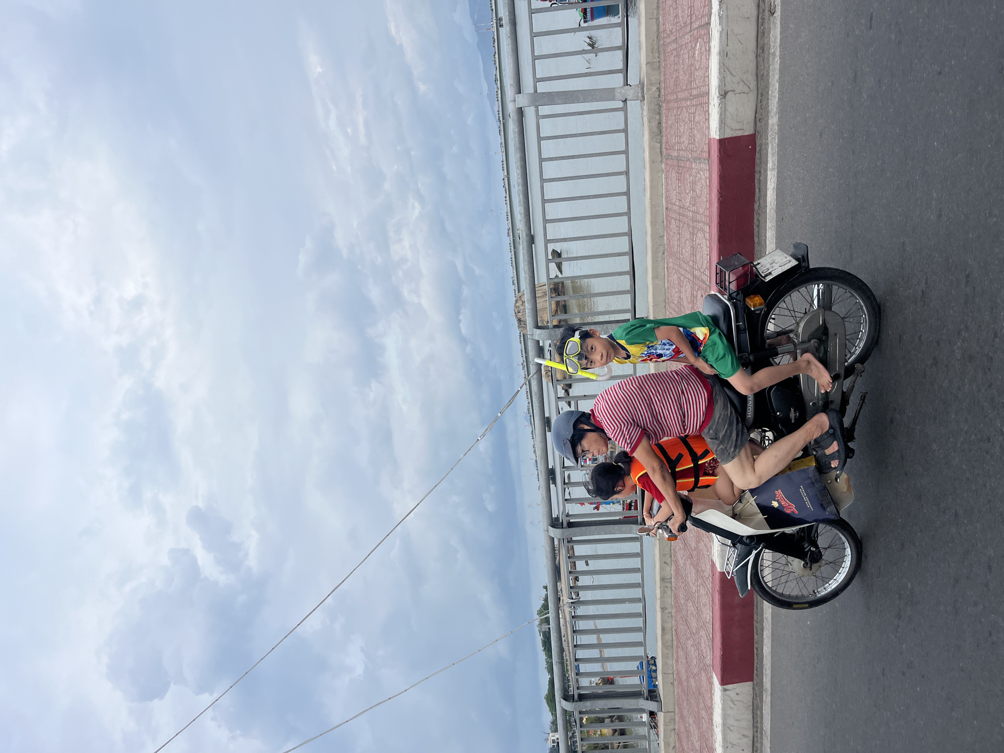 The image depicts a family of three on a motorcycle. The background shows a cloudy sky and a bridge with a metal railing. The bridge overlooks a body of water with distant shoreline visible. The family consists of an adult and two children. The adult wears a striped shirt, helmet, and an orange life vest. One child sits in front, wearing a similar vest. The second child is behind the adult, wearing a green and yellow shirt and goggles. The motorcycle is black and white, parked on an asphalt road. A white curb with red and white stripes runs along the roadside. The location is Bac Nha Trang Ward, Vietnam.