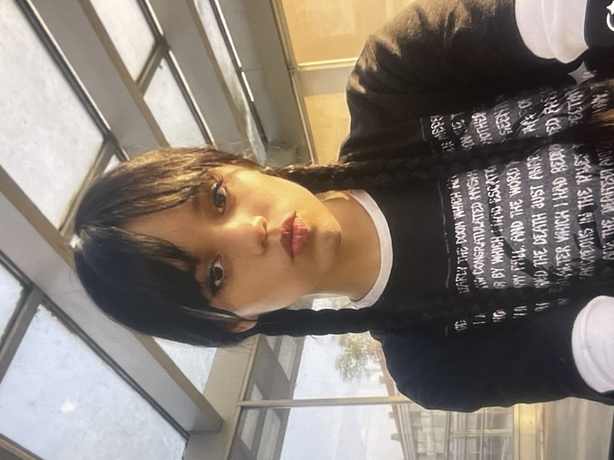 The image displays a young person, likely female, with dark hair styled in two long braids and blunt bangs framing her face. She looks directly at the camera with a neutral to slightly serious expression, featuring dark eyes and lips with a reddish tint.
She is wearing a black long-sleeved t-shirt with a visible white collar, suggesting a layered look or a ringer tee. The shirt is printed with white text, much of which is partially obscured or fragmented due to the angle and creases in the fabric. Visible phrases from the OCR include: "DEARLY THE DOOM WHICH," "IN CONGRATULATED MYSHO," "BY WHICH TIHID ESCAT," "MOTHER VAY PILL AND THE WORDSEEY," "AY AND THE DEATH JUST ANDE APE OF," and "SLTER WHICH I HAD REDIO TED THE ANDRCONS IN THE TRUETY PECTIN ARE THE DERITEIT AND." The text also includes "CD" and "11."
The background reveals large windows with metal frames, through which bright, diffused daylight is visible, indicating daytime. The setting appears to be an indoor or semi-enclosed space, possibly a modern building or covered walkway, with reflections on the glass. The person is posing for a portrait, likely a selfie. The location context provided is Poltava, Ukraine.