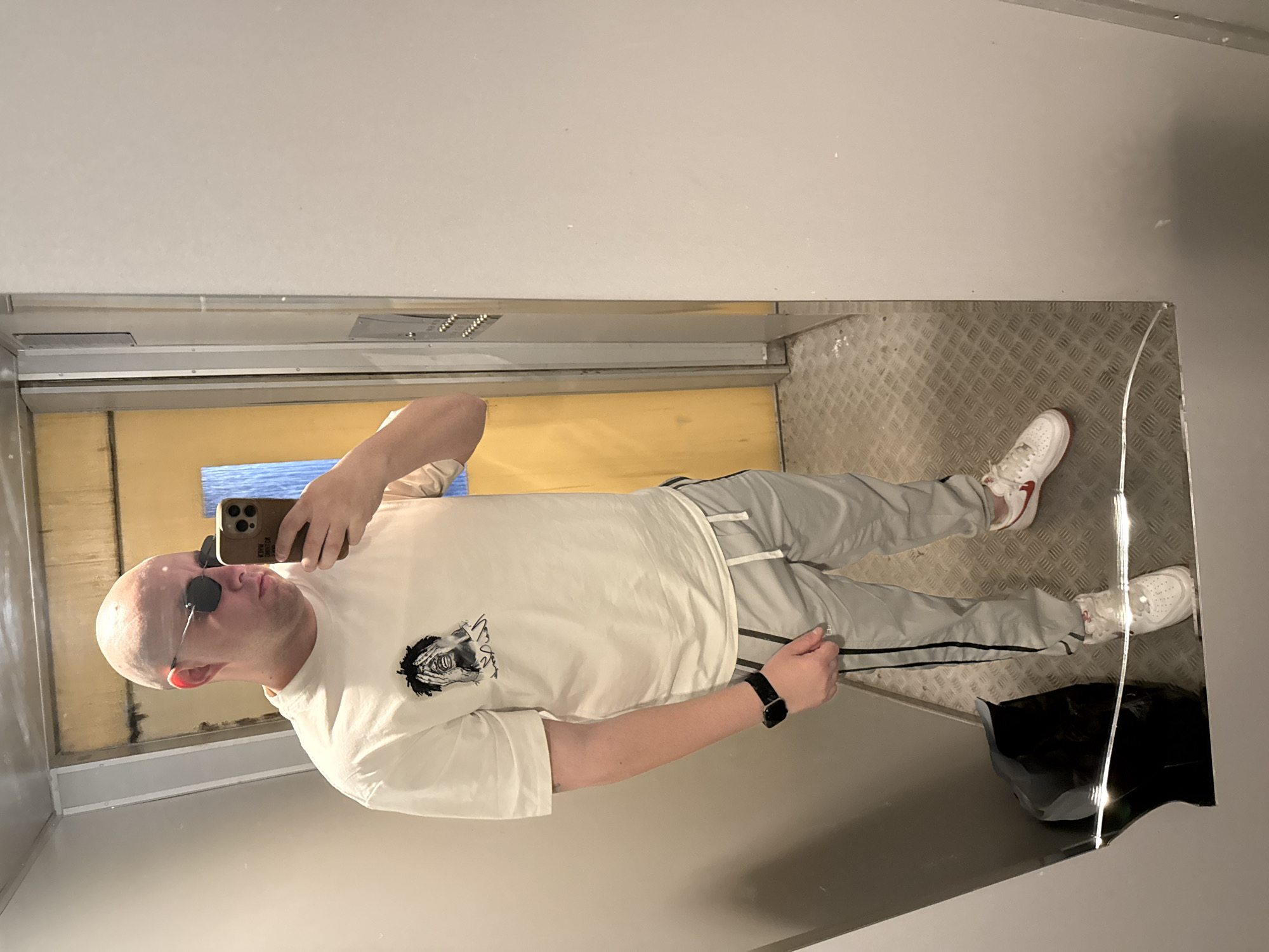 A fair-skinned, bald man with sunglasses is taking a selfie in an enclosed space with reflective walls, likely an elevator. He is wearing a white t-shirt with a graphic print, light gray track pants, and white sneakers with red accents. A black wristwatch is visible on his left wrist. The floor of the enclosure is textured metal. Behind him, a yellow wall is visible, with a strip of blue running horizontally. A black garbage bag is in the bottom right corner of the reflective space. The photograph's orientation is rotated. It is not possible to ascertain the exact location in Pleven, Bulgaria, from the provided image.
