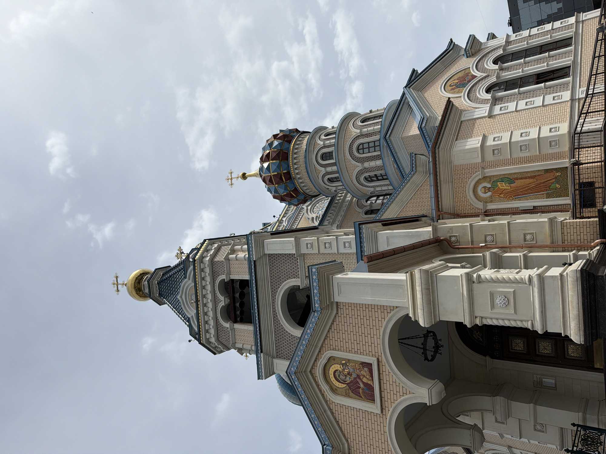 The image shows the exterior of St. Michael's Cathedral in Izhevsk, Russia. The cathedral features elaborate brickwork, arched windows, mosaics depicting religious figures, and a distinctive onion dome topped with a golden cross. The structure also includes a bell tower. The sky is overcast. The building is shot from a low angle.
